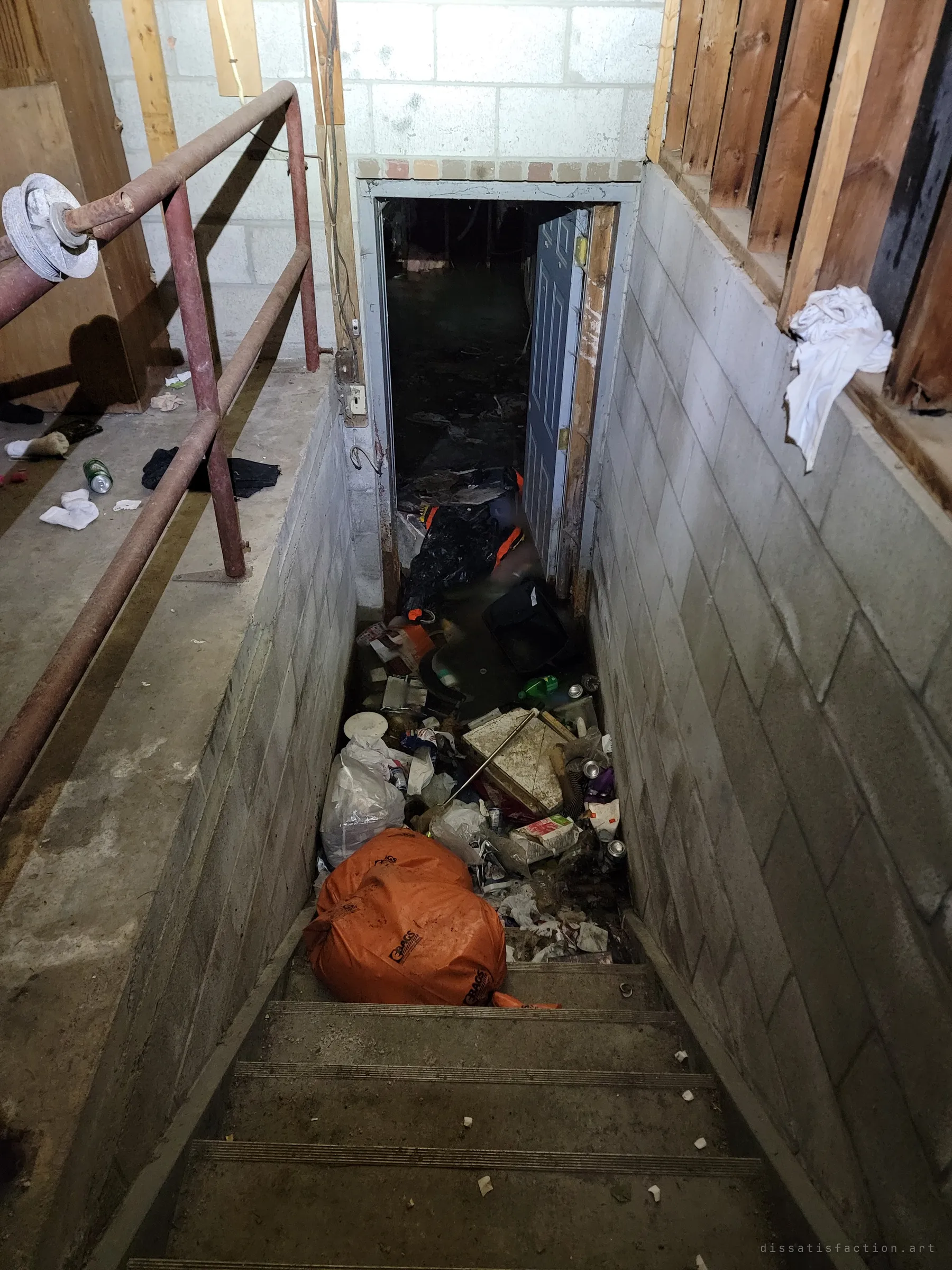 Looking down the concrete-block stairwell from the garage into the basement, the steps choked with garbage bags, cans and debris. The basement door hangs open onto darkness.