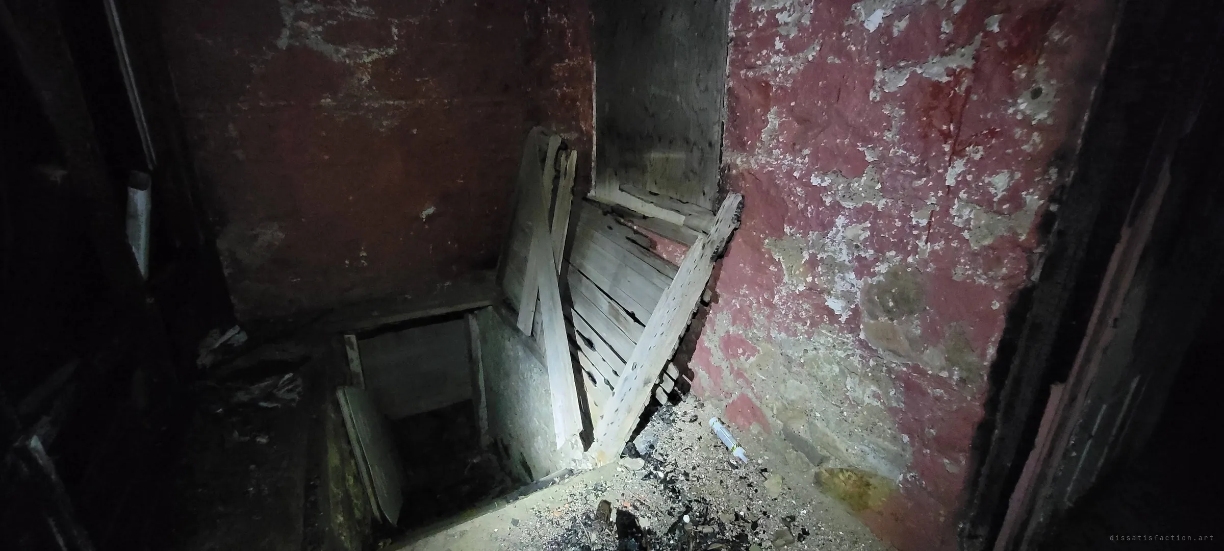 A trapdoor opening in a scorched floor drops into a cellar, beside boarded-up planks and flaking red plaster walls. The fire damage and makeshift barricade show how far the house has fallen.