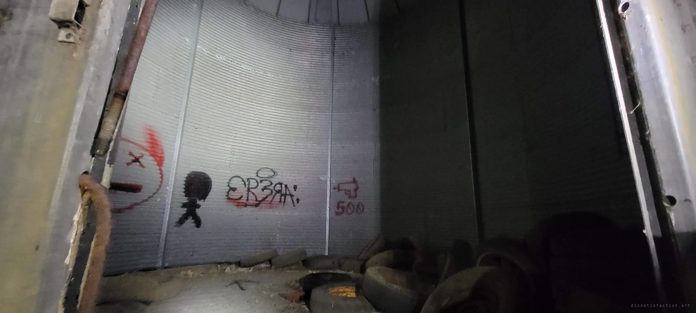 Looking into a corrugated steel grain bin through its hatch, with spray-painted graffiti on the curved wall and old tyres piled on the floor. The farm's storage has become a canvas for visitors.
