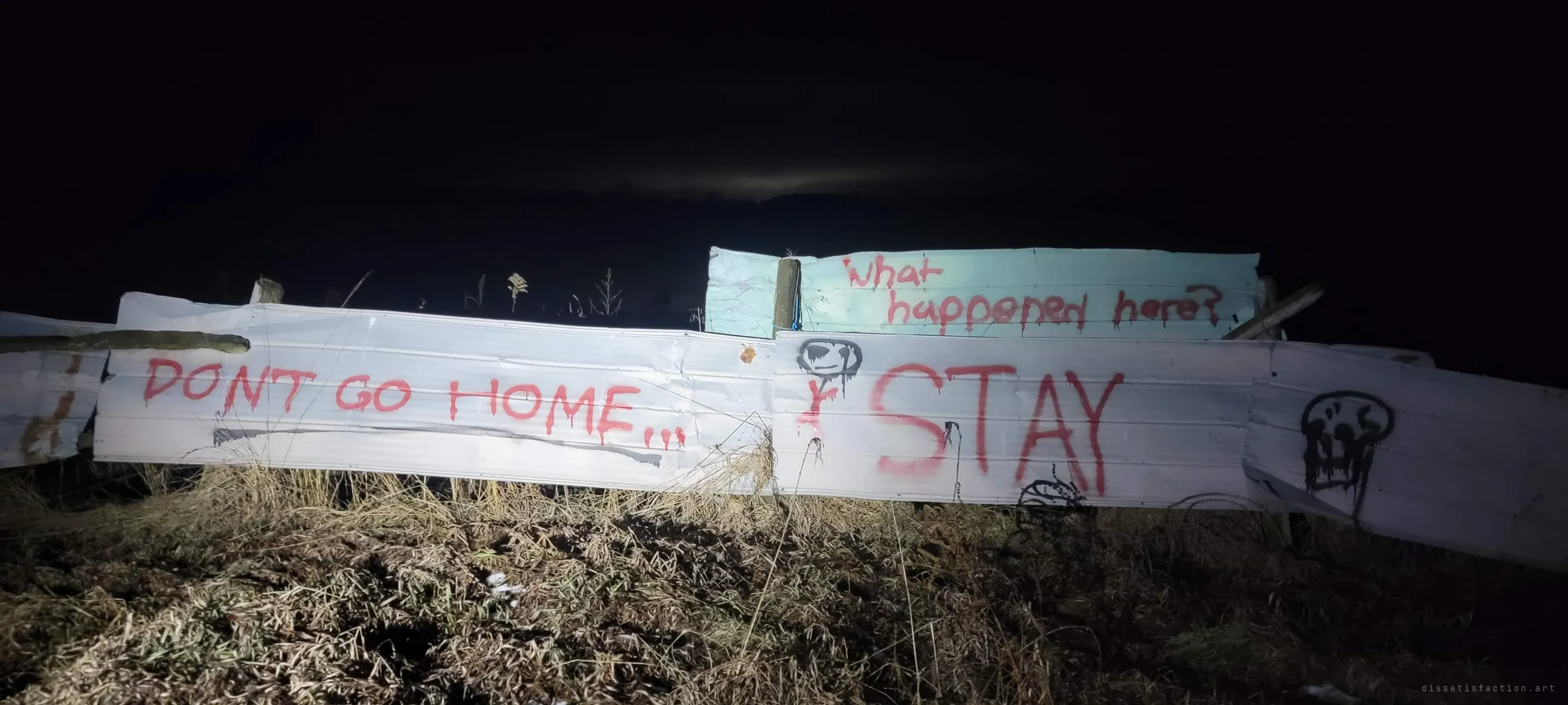 Flashlit sheet-metal fence panels at night, covered in dripping red painted warnings and cartoon skulls. It sets the ominous tone of the property from the outside.