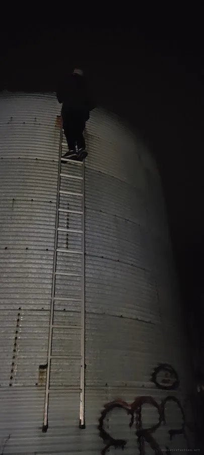 A figure stands at the top of a tall ladder bolted to the side of a corrugated steel silo in the dark. It shows the height and risk of the climb.