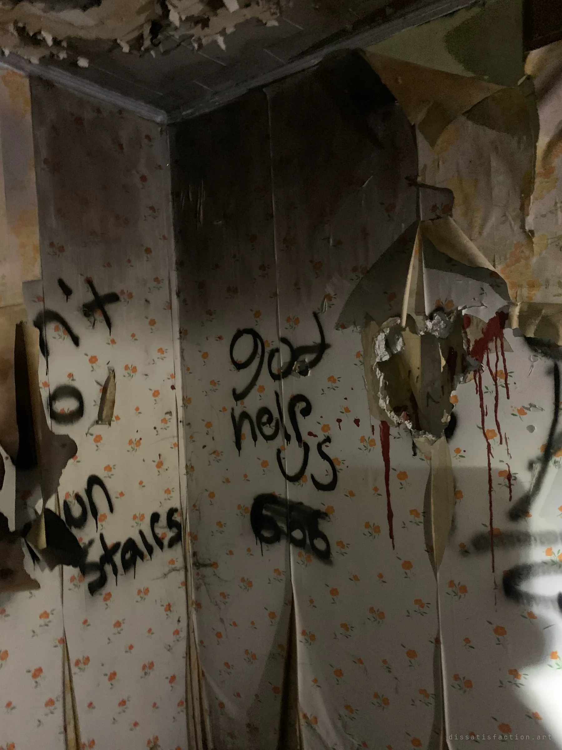 A corner of a room with peeling floral wallpaper, black spray-painted messages and red drips running down the wall. The graffiti turns the old bedroom into something out of a horror film.