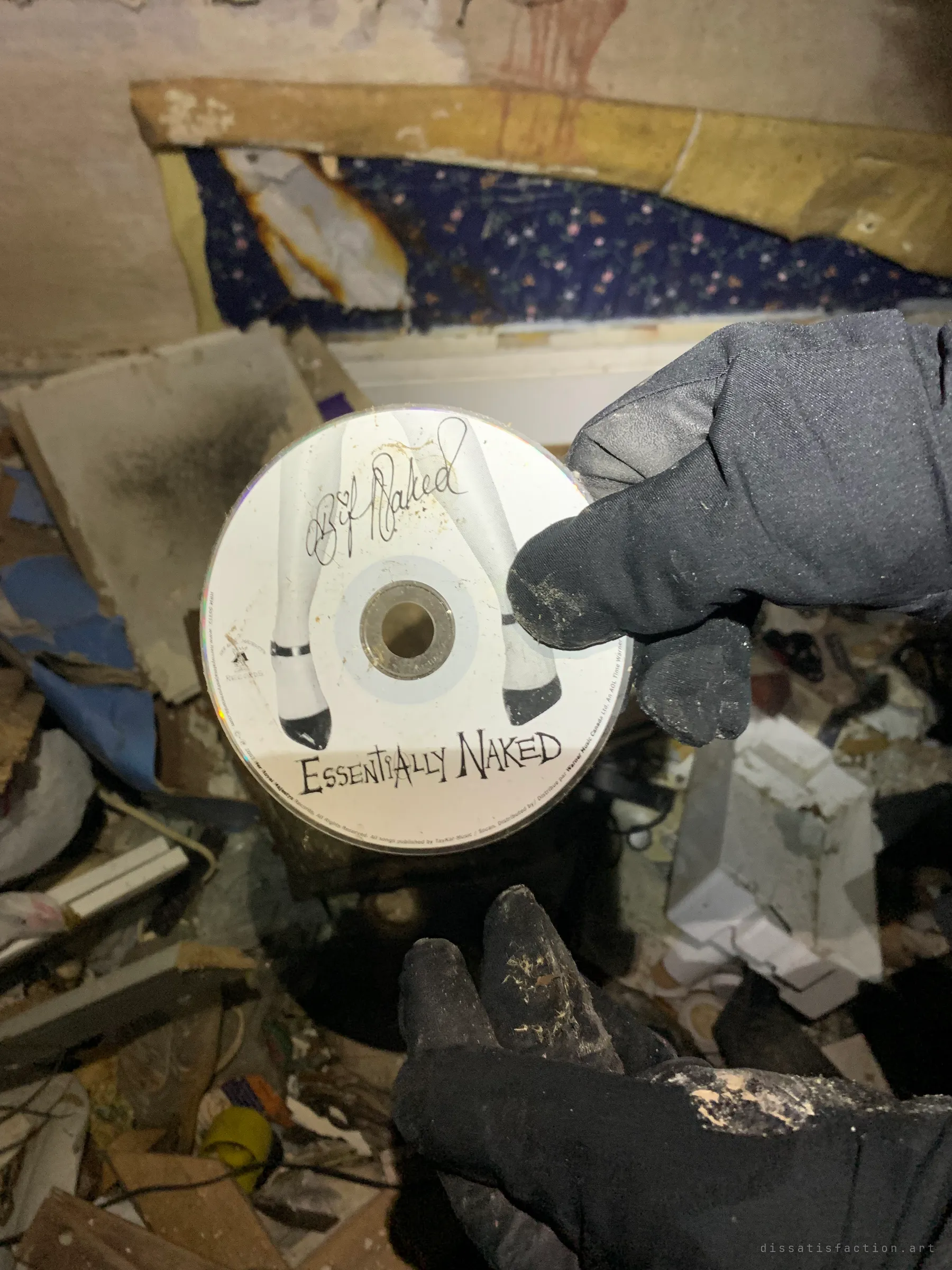 A gloved hand holds up a dusty music CD found among the debris of a burnt room. A small personal leftover from whoever lived here.