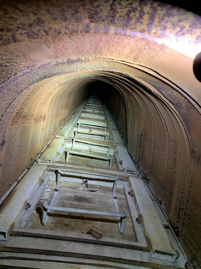 A narrow rusted metal chute with a ladder of rungs running down into darkness. The tight tunnel shows the farm's old grain-handling machinery.