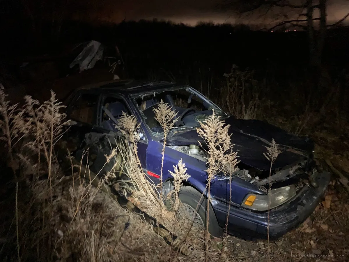 A wrecked blue sedan with a crushed hood sits abandoned in tall dry weeds at night. It has been left to rot along with the rest of the farm.