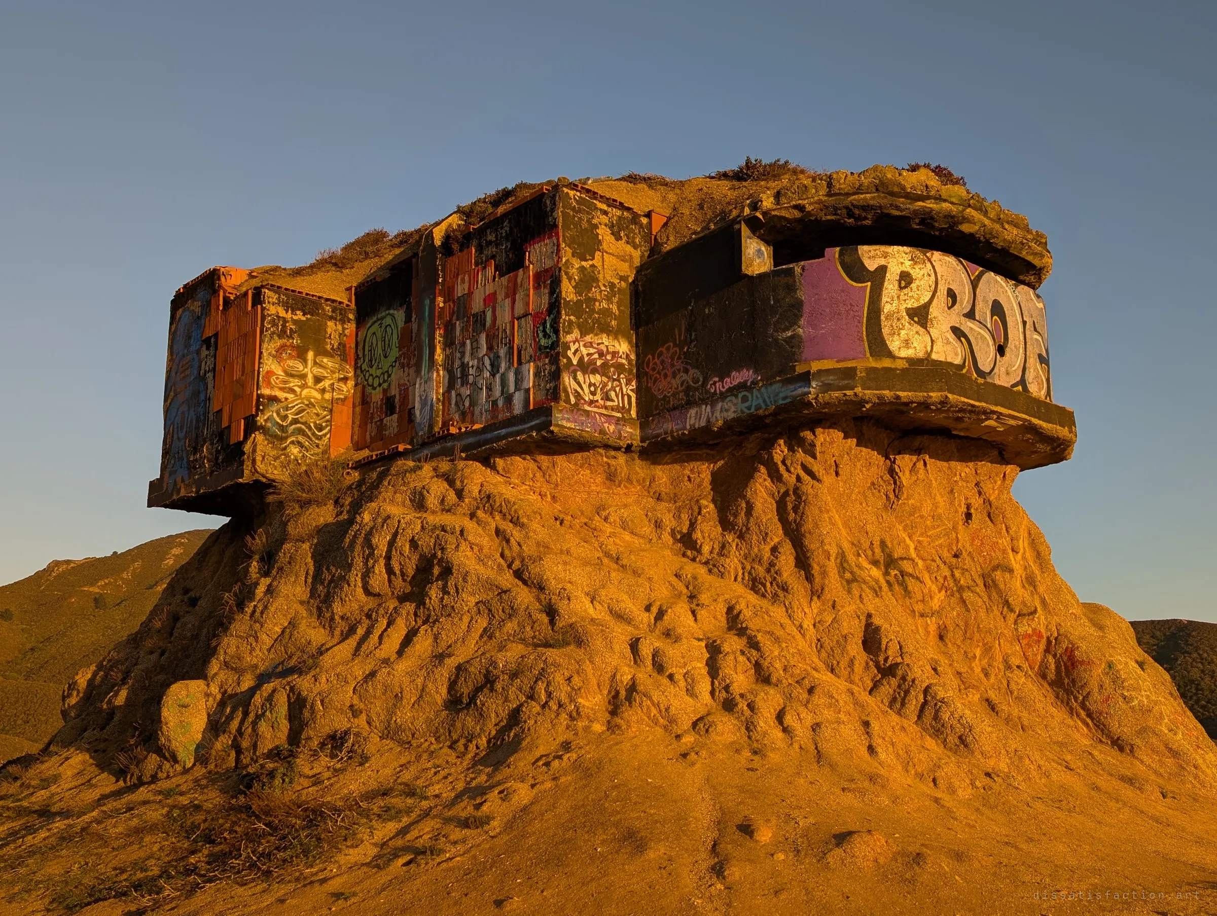 A graffiti-covered concrete bunker sits on top of a crumbling, eroded rock pillar, glowing orange in low evening sun. The ground has worn away beneath it, leaving the structure overhanging its pedestal.