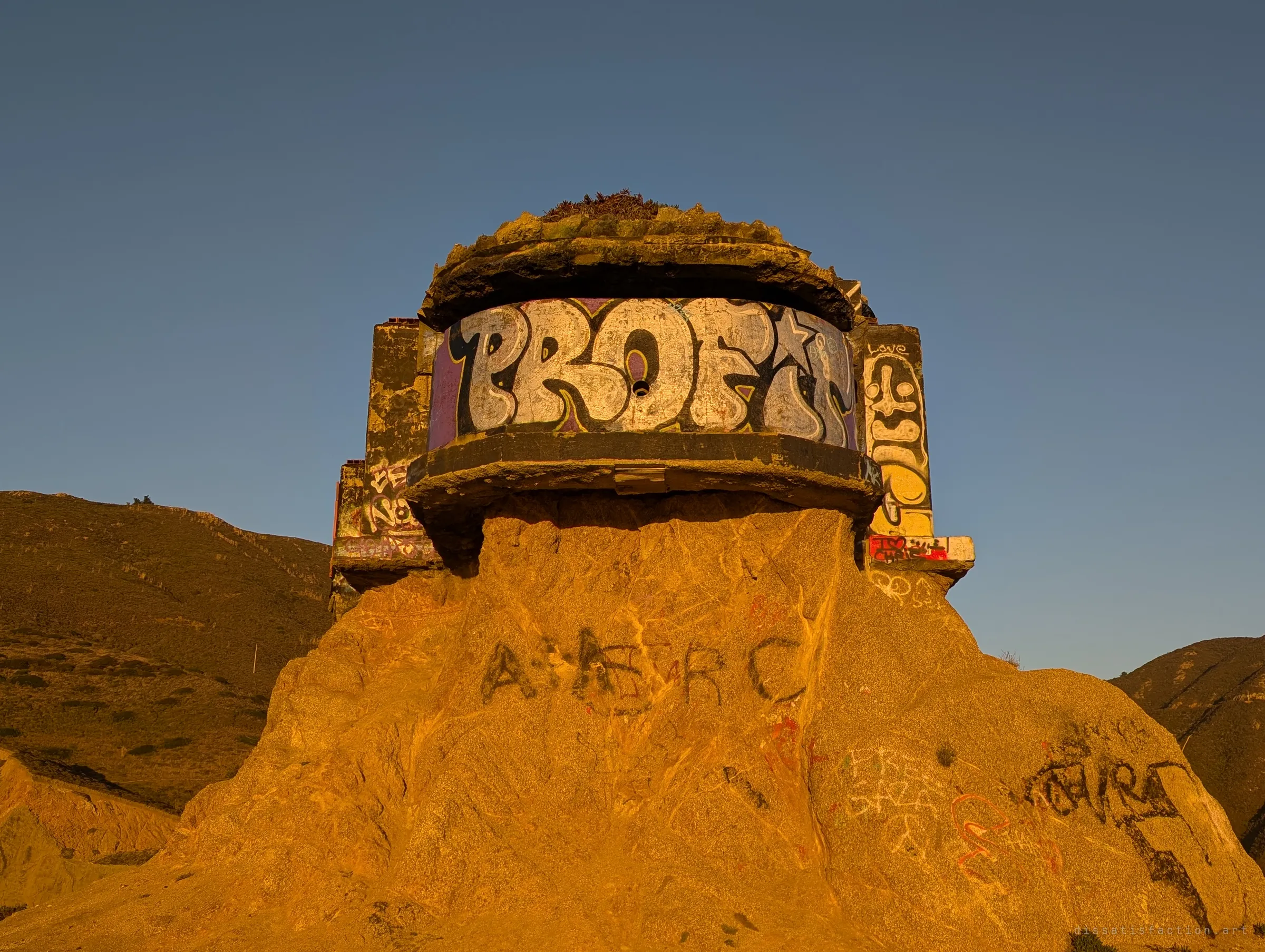 Looking up at the rounded front of the bunker, its curved wall covered in a large graffiti piece, resting on a sunlit rock stump carved with scratched tags. Dry hills rise on either side under a clear sky.