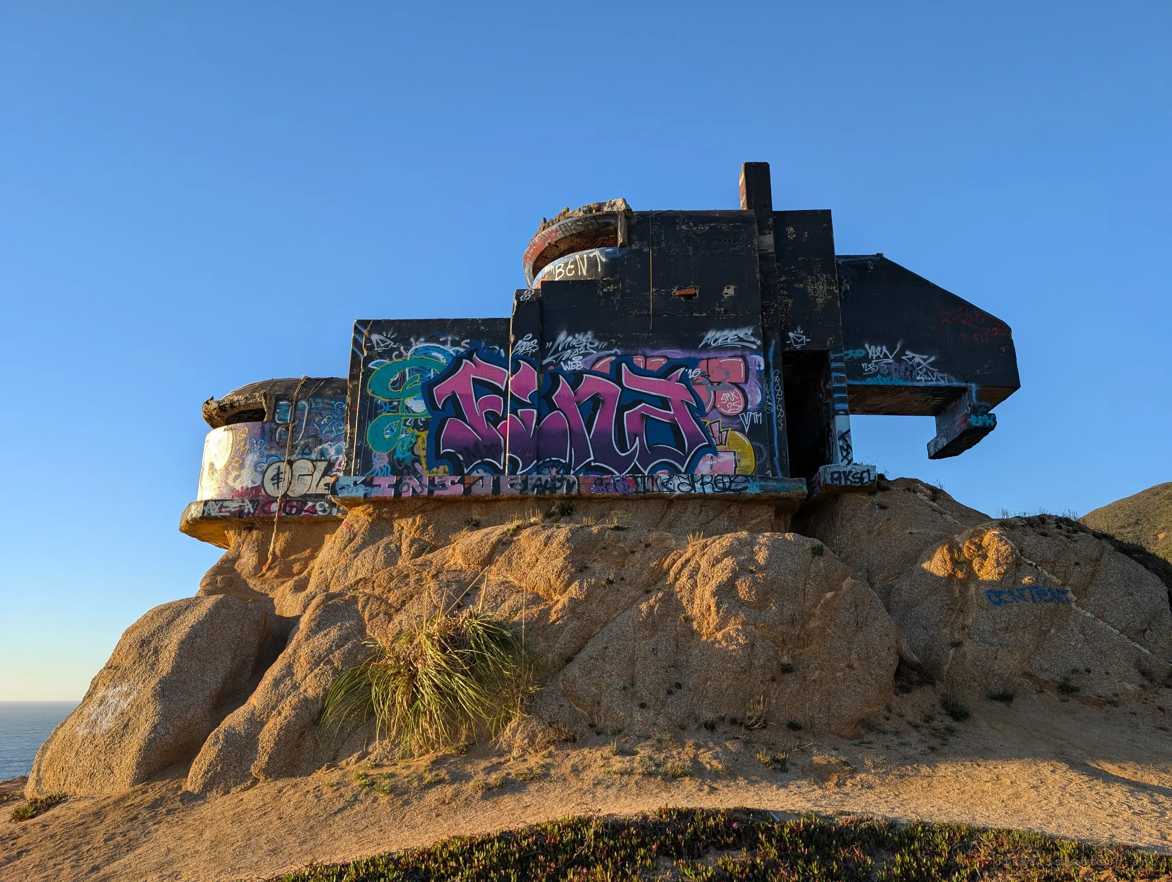 The long side of the bunker, a stack of dark concrete blocks with a cantilevered arm, painted with bright pink and blue graffiti and balanced on rounded boulders. A sliver of ocean shows at the far left.