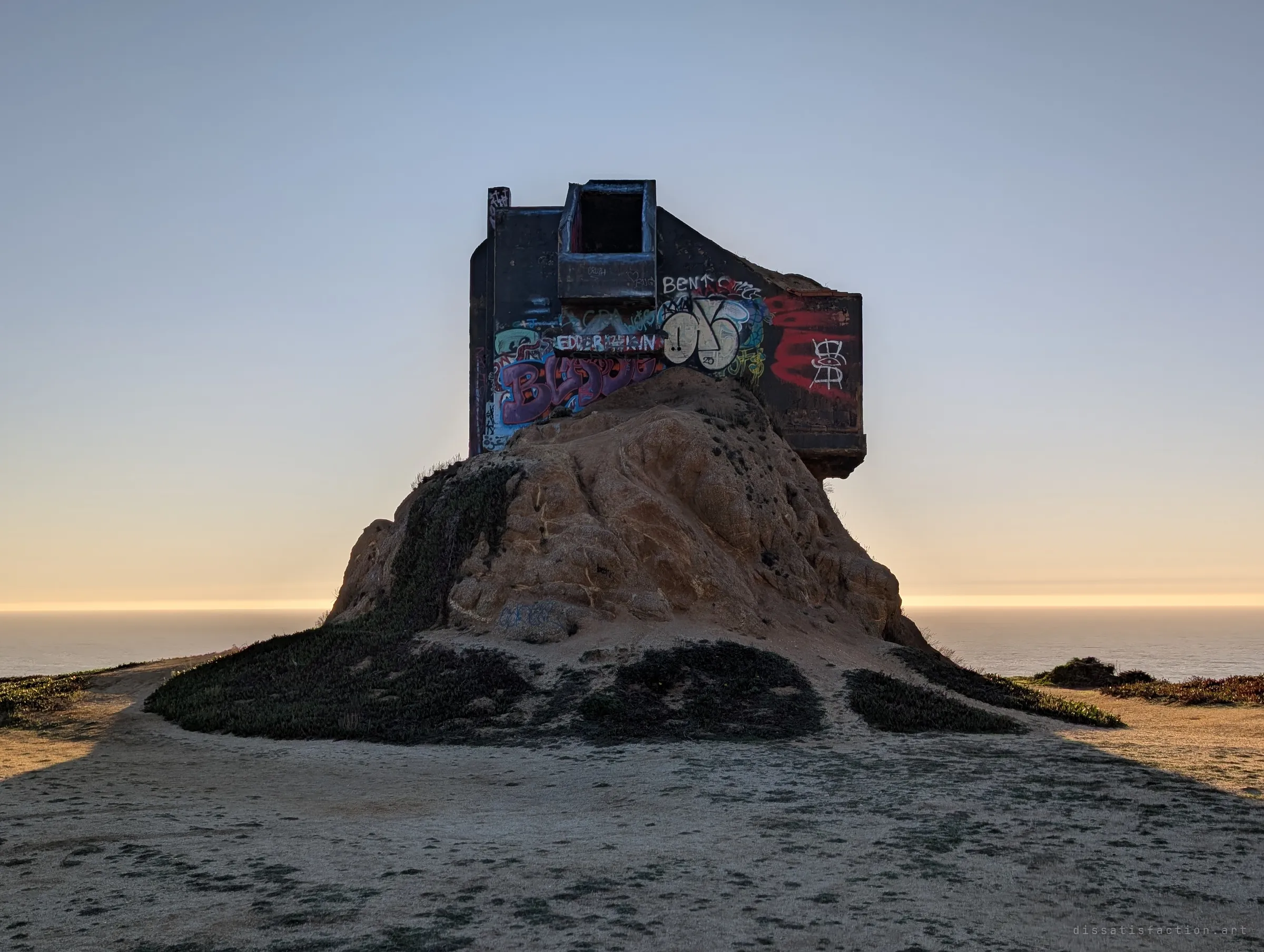 The bunker seen from behind at dusk, a dark graffiti-covered box on a lone rock mound with a flat ocean horizon glowing on either side. A worn dirt clearing leads up to it.
