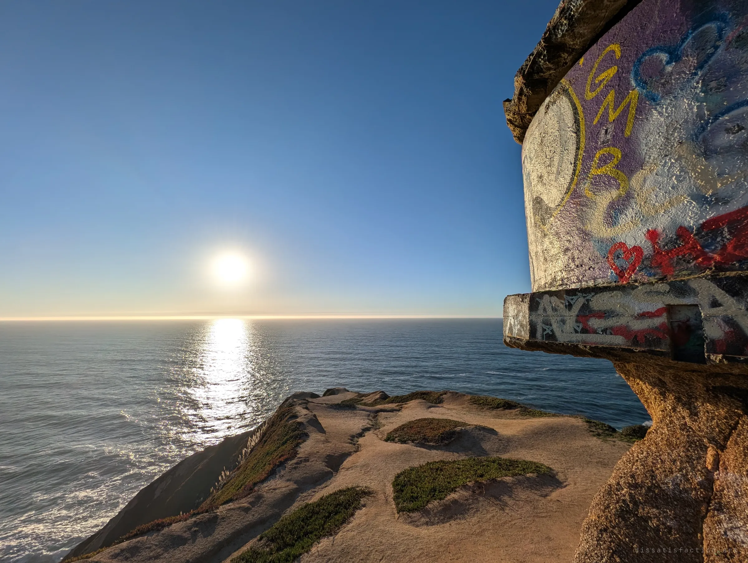 A wide view from the bunker's edge: its painted curved wall juts in from the right over a sandy clifftop, with the low sun shining over the open ocean. It shows how the structure hangs above the coast.