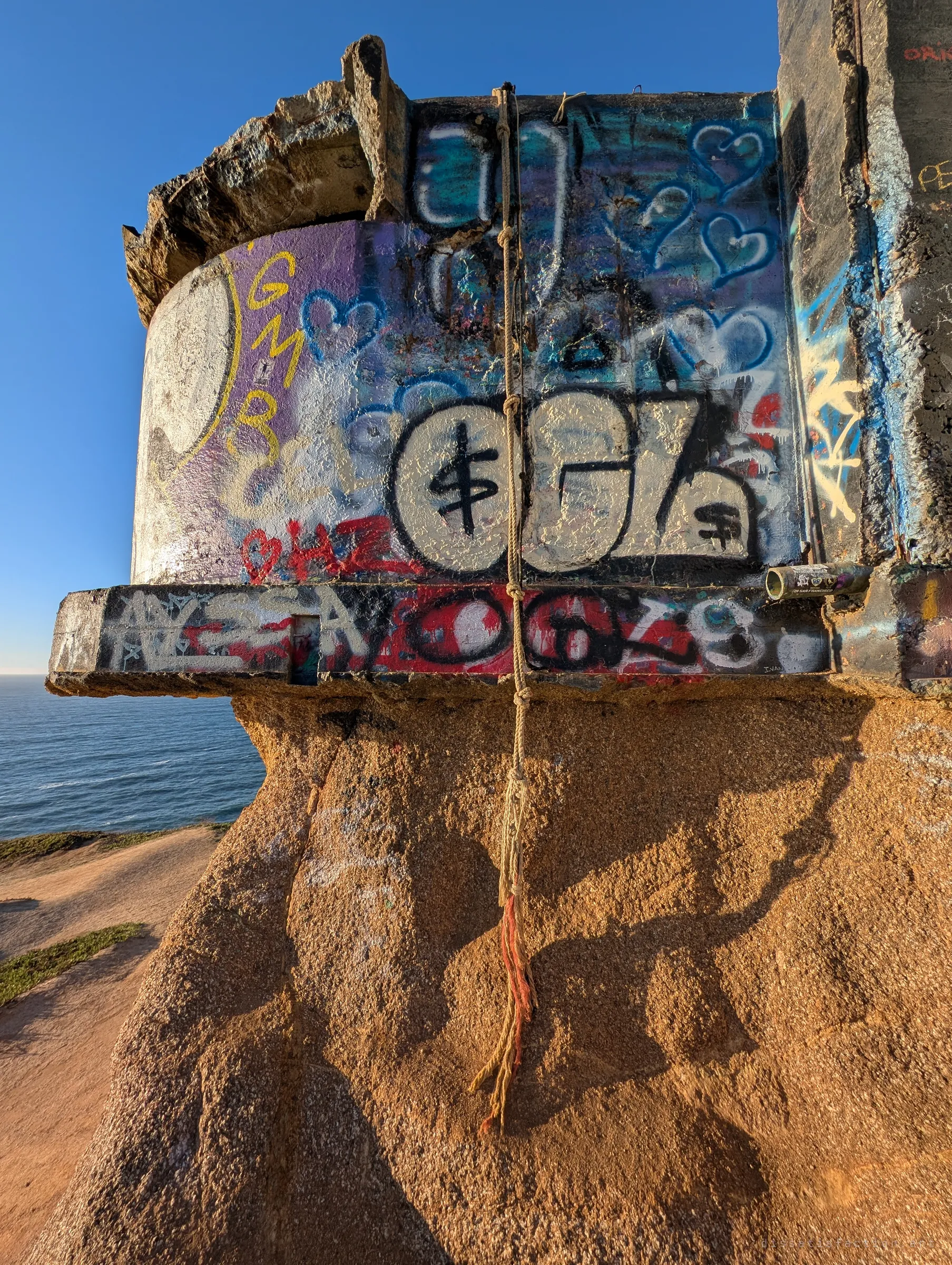 Close up of one corner of the bunker, layered with hearts, tags and throw-ups, with a frayed rope hanging down the eroded rock below. The ocean sits far beneath on the left.