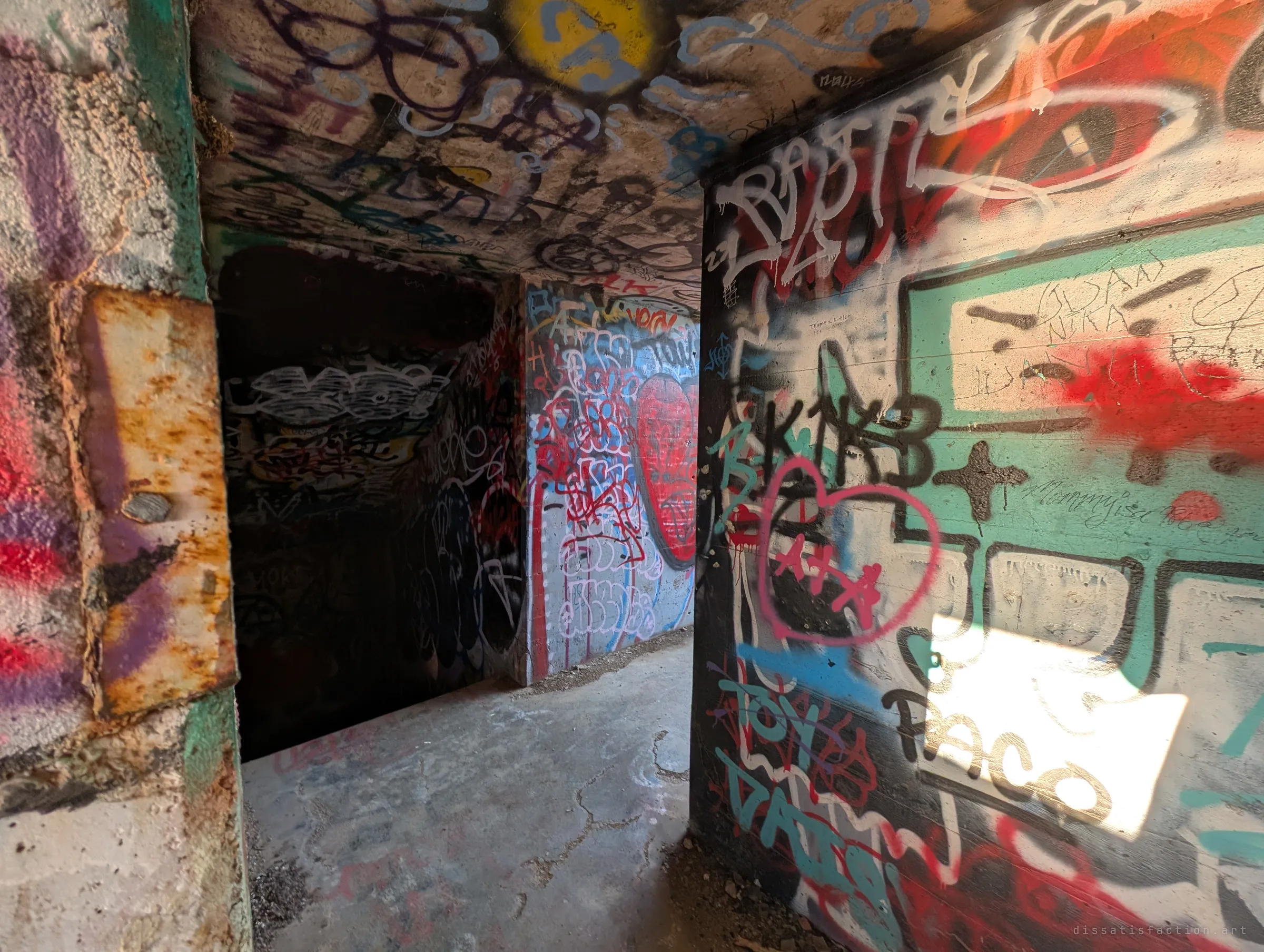 Ultra-wide view through a doorway with a rusted steel frame into dim graffiti-covered chambers, a patch of hard sunlight hitting the right wall. The thick poured concrete shows the bunker's military build.