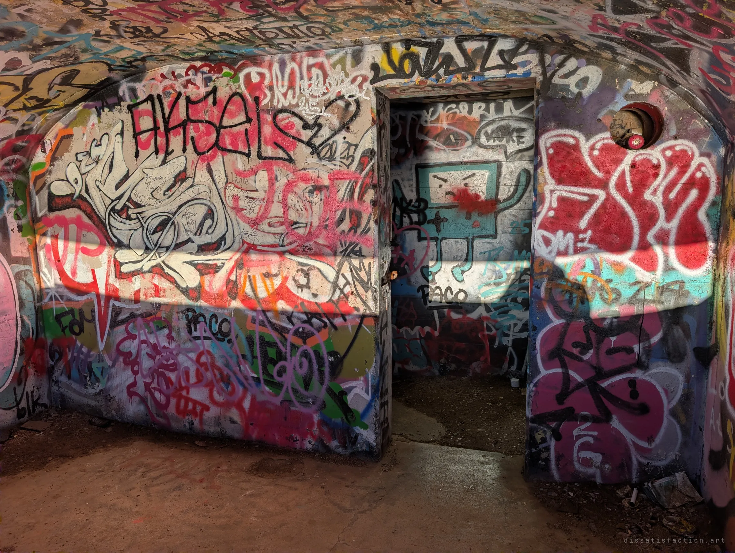 A vaulted concrete room covered wall to ceiling in tags, with a cartoon robot painted inside a small doorway and a band of low sunlight cutting across. A round vent hole sits high on the right wall.