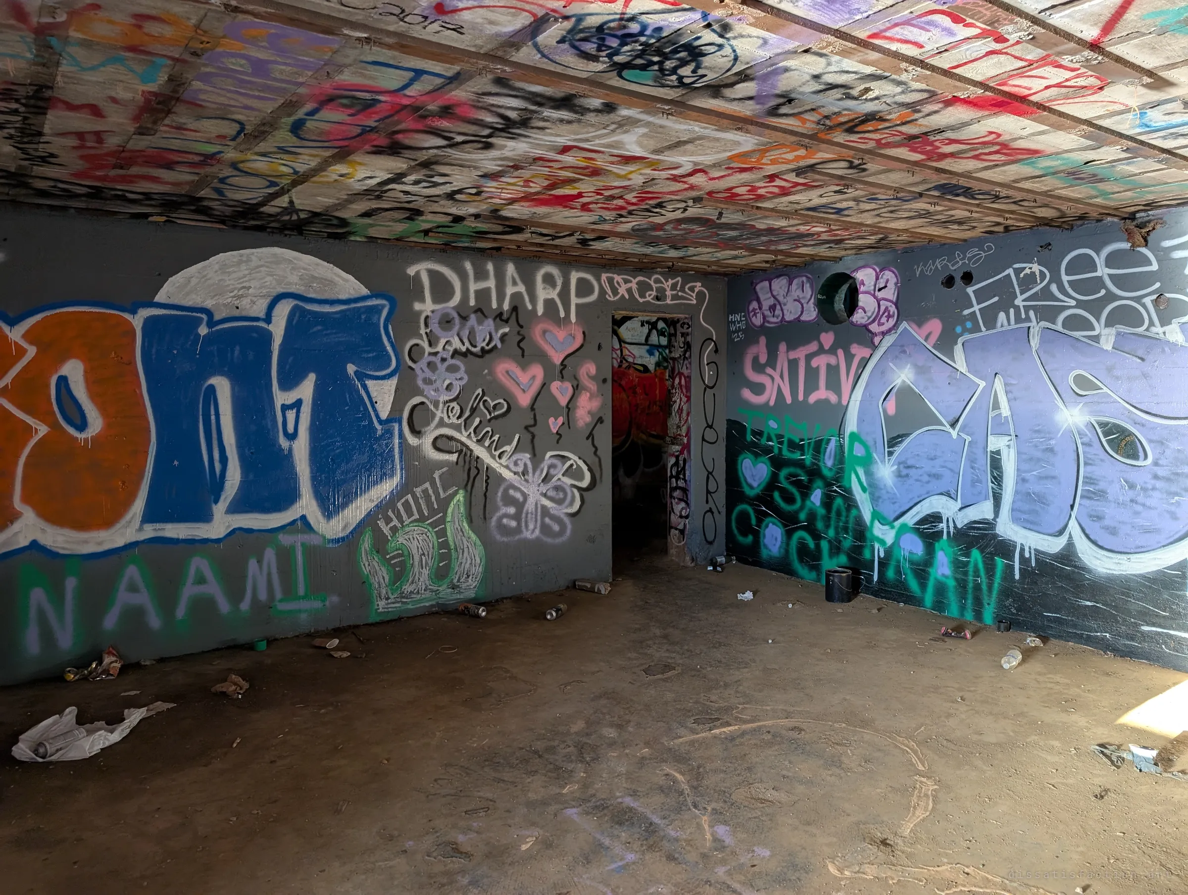 A larger rectangular chamber with a board-formed concrete ceiling, big painted pieces on the grey walls and spray cans scattered across the dirt floor. A doorway at the back leads deeper into the structure.