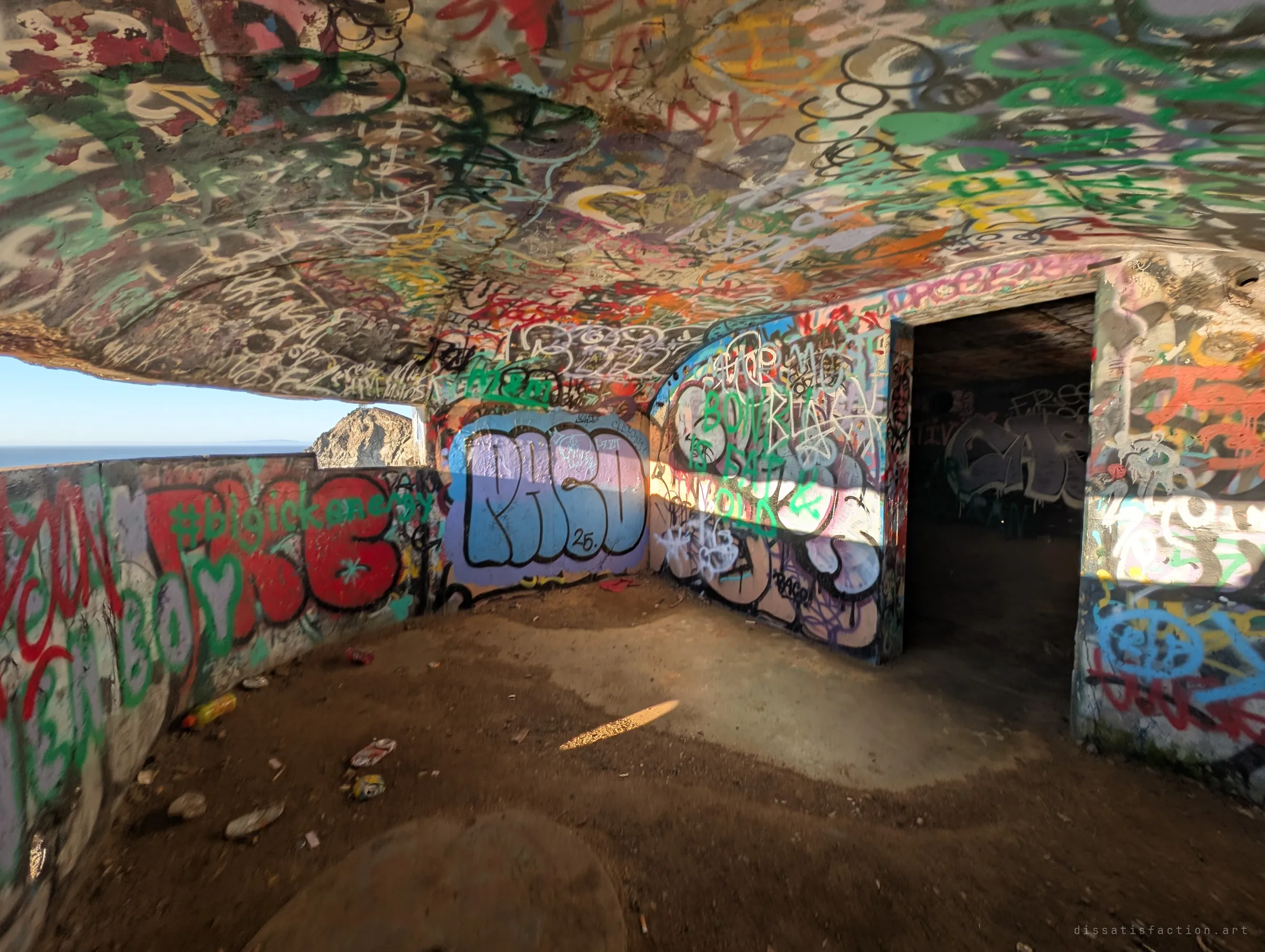 An ultra-wide look at a graffiti-soaked observation room, its long horizontal opening letting in daylight and a glimpse of sea. Tags cover every surface including the curved ceiling.