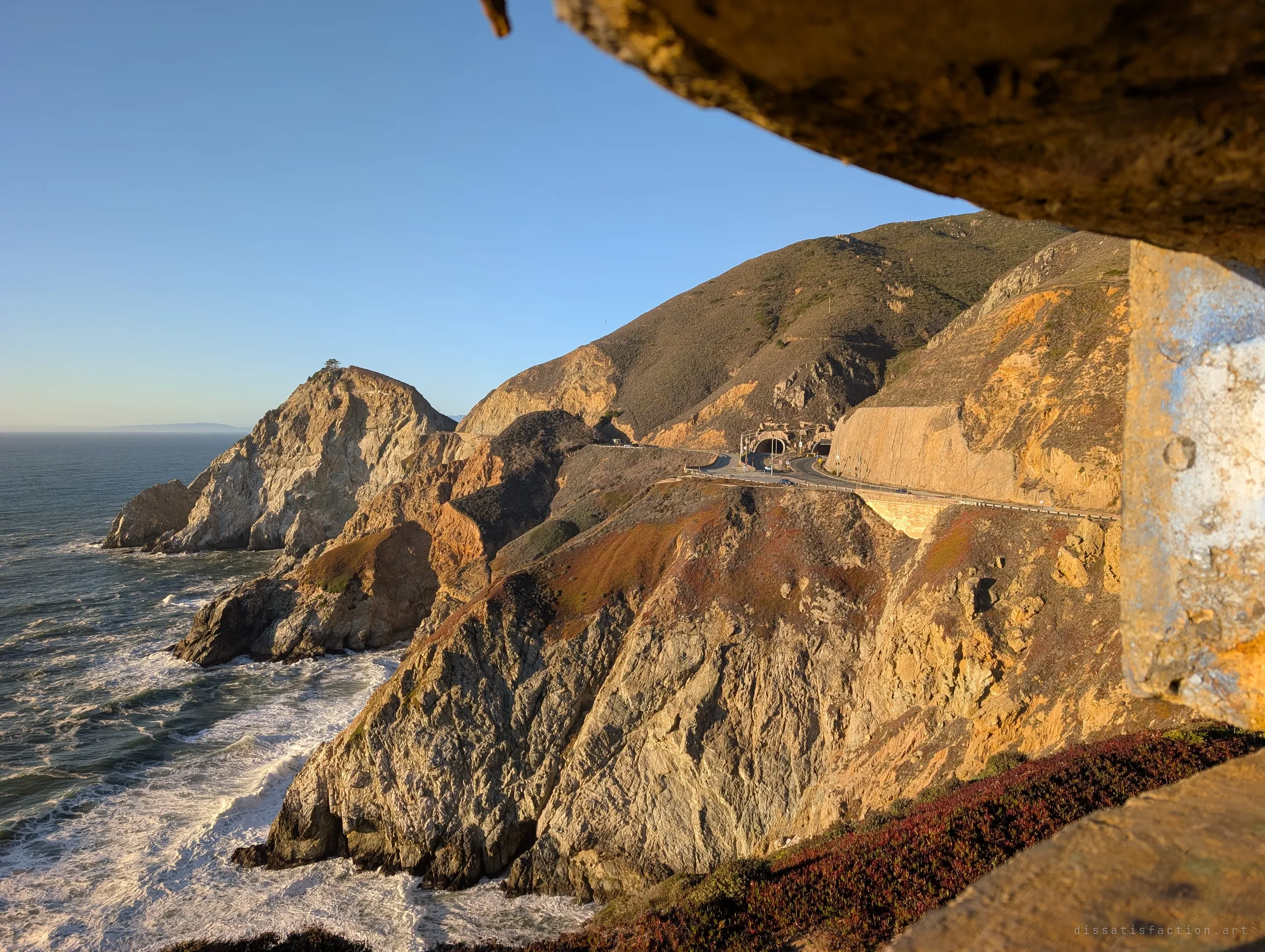 Looking out from under a concrete lip over steep sunlit cliffs, crashing surf and a coastal highway cut into the hillside. It shows why the bunker sits where it does: a commanding watch over the sea approach.