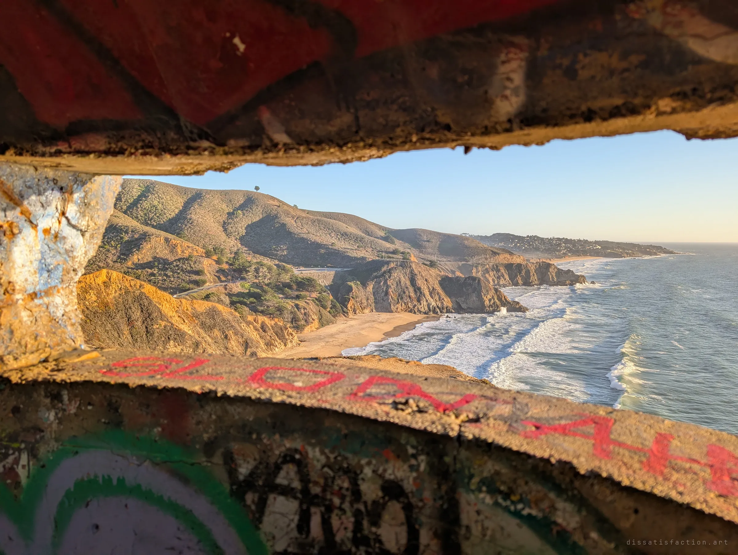 The ocean and a long curving coastline framed between the rough concrete lips of a gun slit, with graffiti on the sill in the foreground. The firing port turns the view into a lookout.