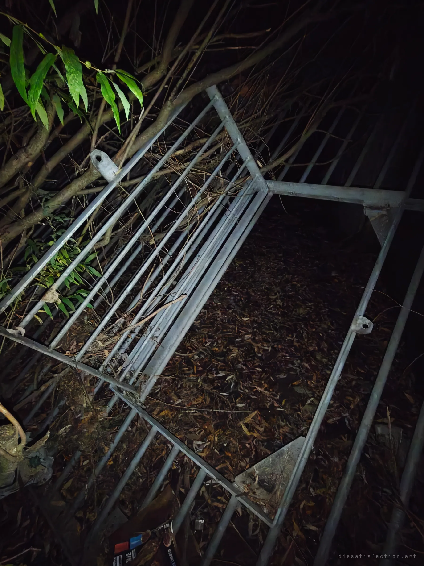 A galvanised steel trash rack lies tipped on its side in the undergrowth, its bars buried in fallen leaves and branches. It shows the grate that once guarded the drain's mouth, now pulled loose and left in the brush.