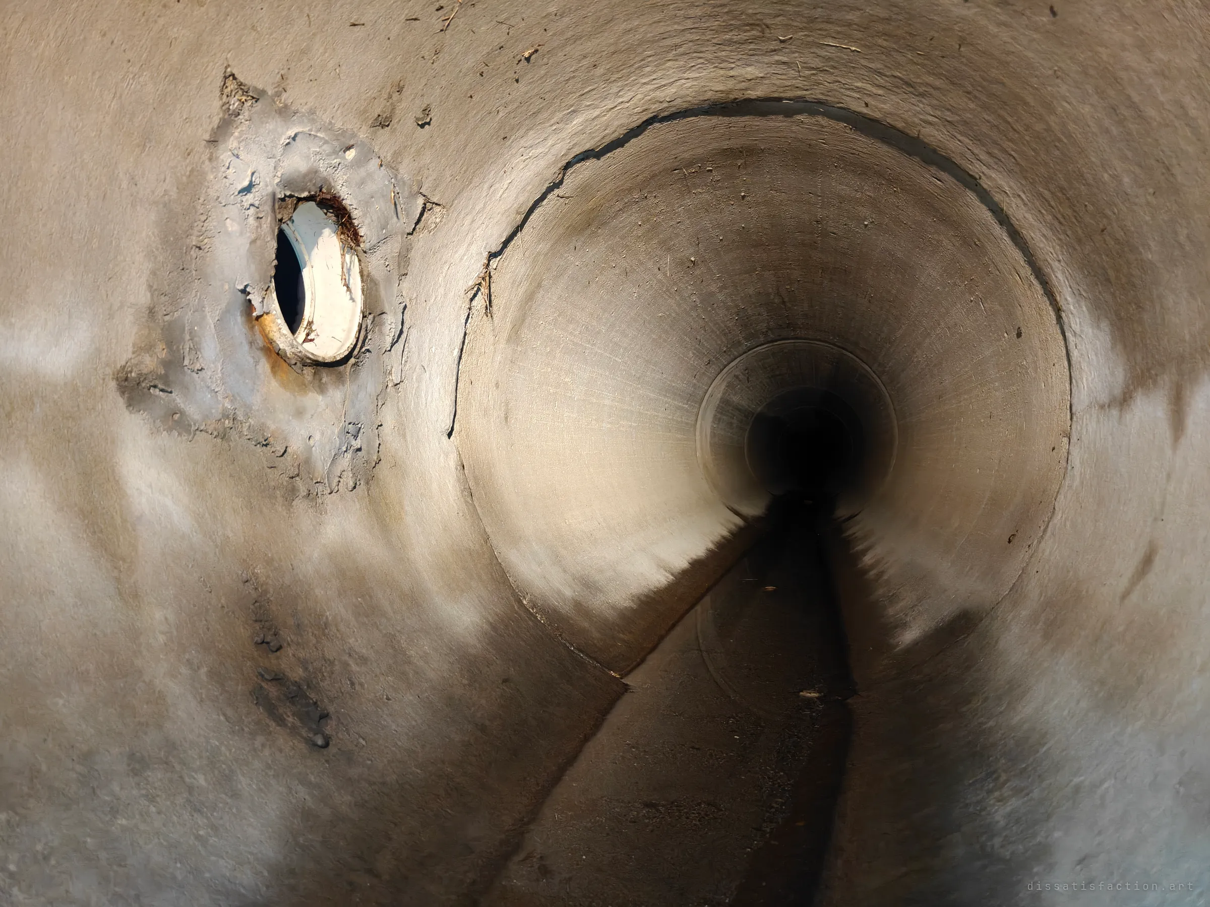 Looking down a round concrete storm pipe that narrows to a smaller barrel at its end. On the left, a white plastic lateral pipe has been patched roughly into the wall, and a low channel of water runs along the floor into the dark.