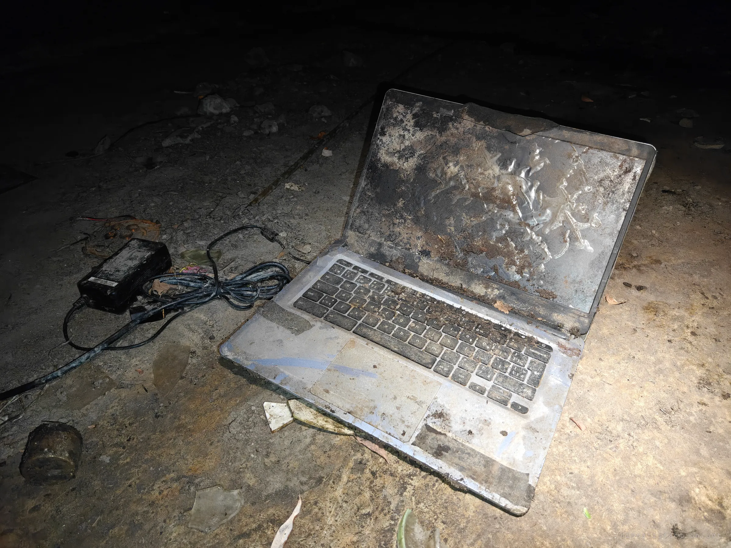 A ruined laptop lies open on the concrete drain floor, its screen caked in dried mud and its keyboard full of silt, with the charger and tangled cable beside it. Something somebody once worked on, washed down here and left to the dark.