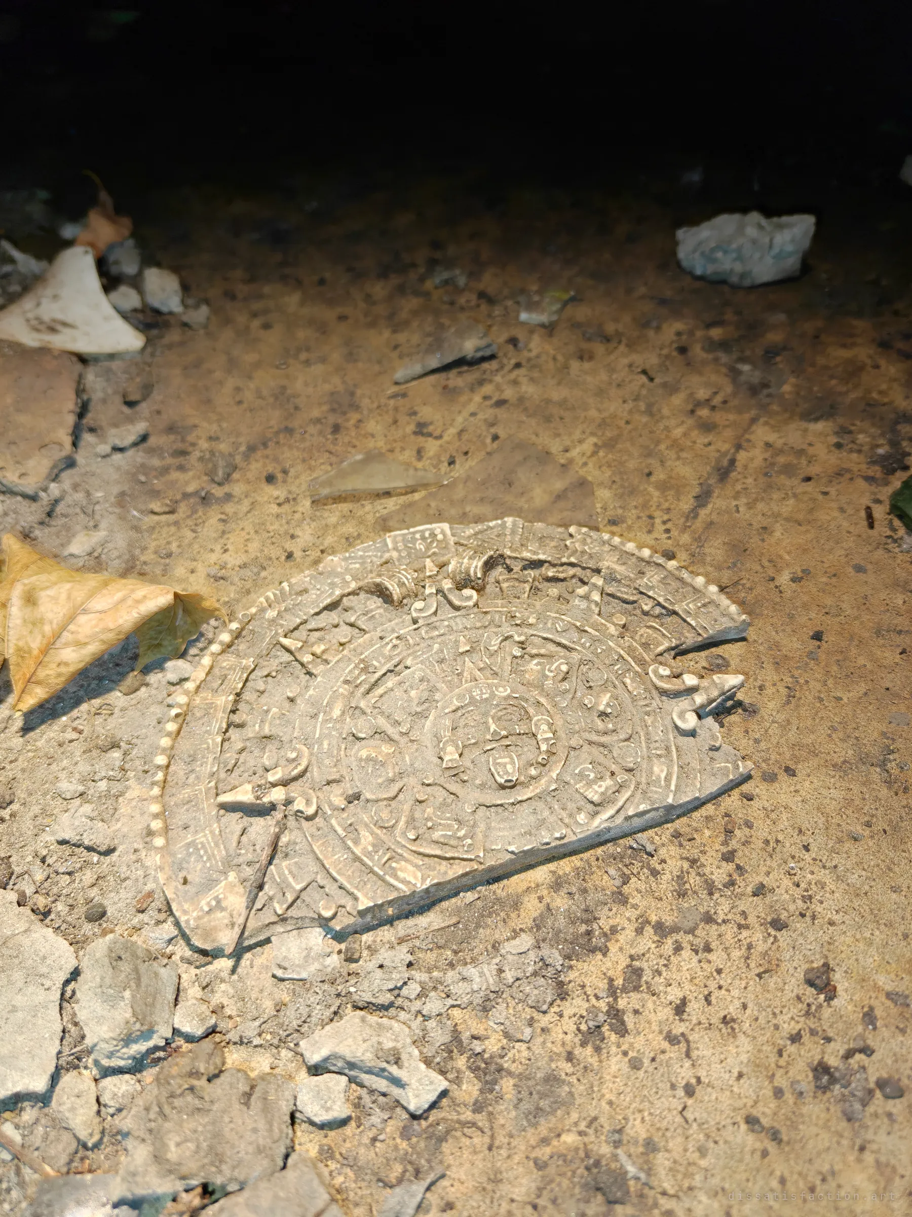 A broken plaster replica of the Aztec sun stone lies on the drain floor among dust, concrete chunks, glass shards and a dried leaf, lit by a flashlight. It is an odd bit of debris washed or dumped into the pipe.