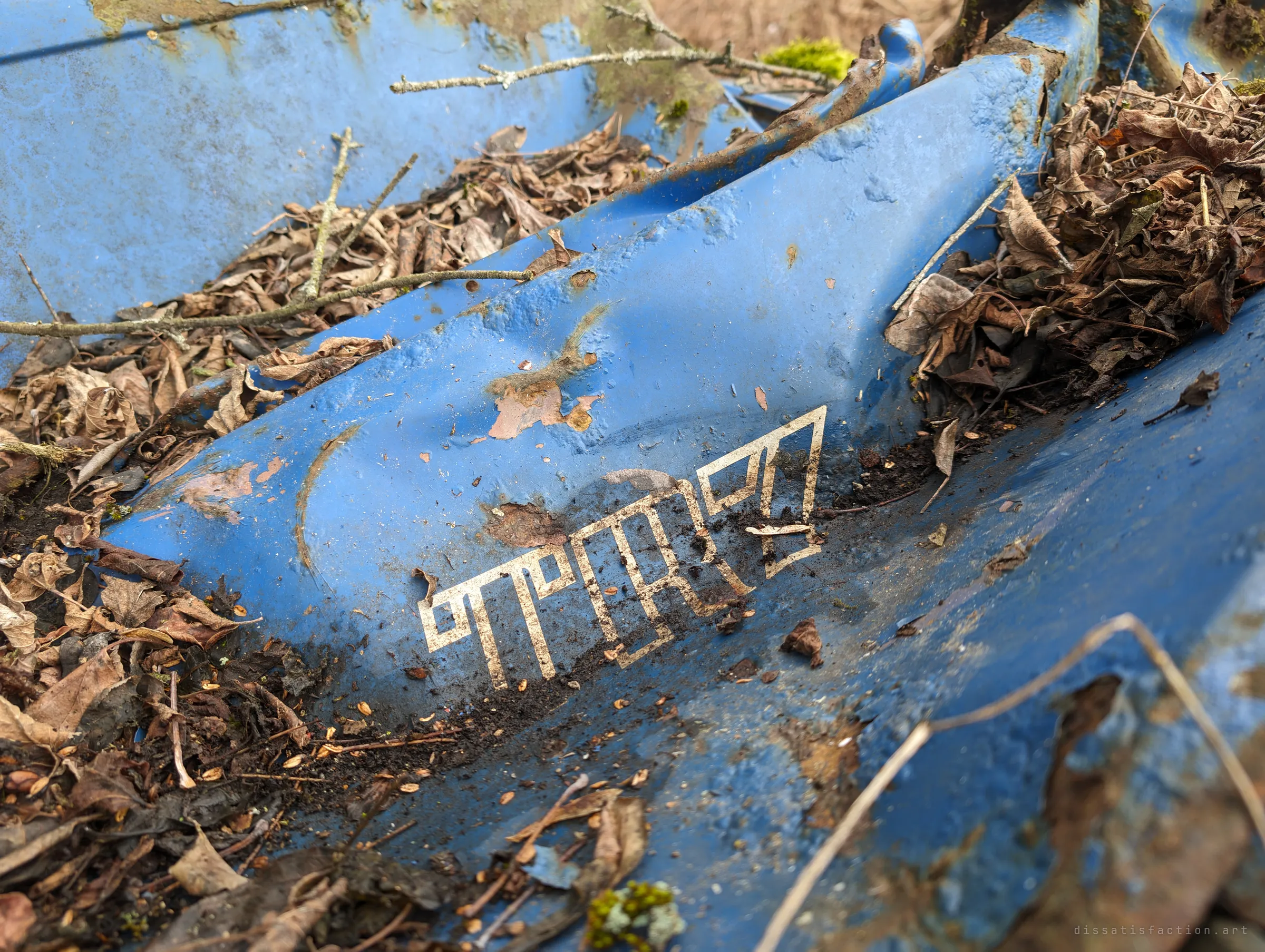 A faded model badge in blocky gold letters on a dented blue panel half buried in dry leaves. The worn lettering is one of the last clues to what the car once was.
