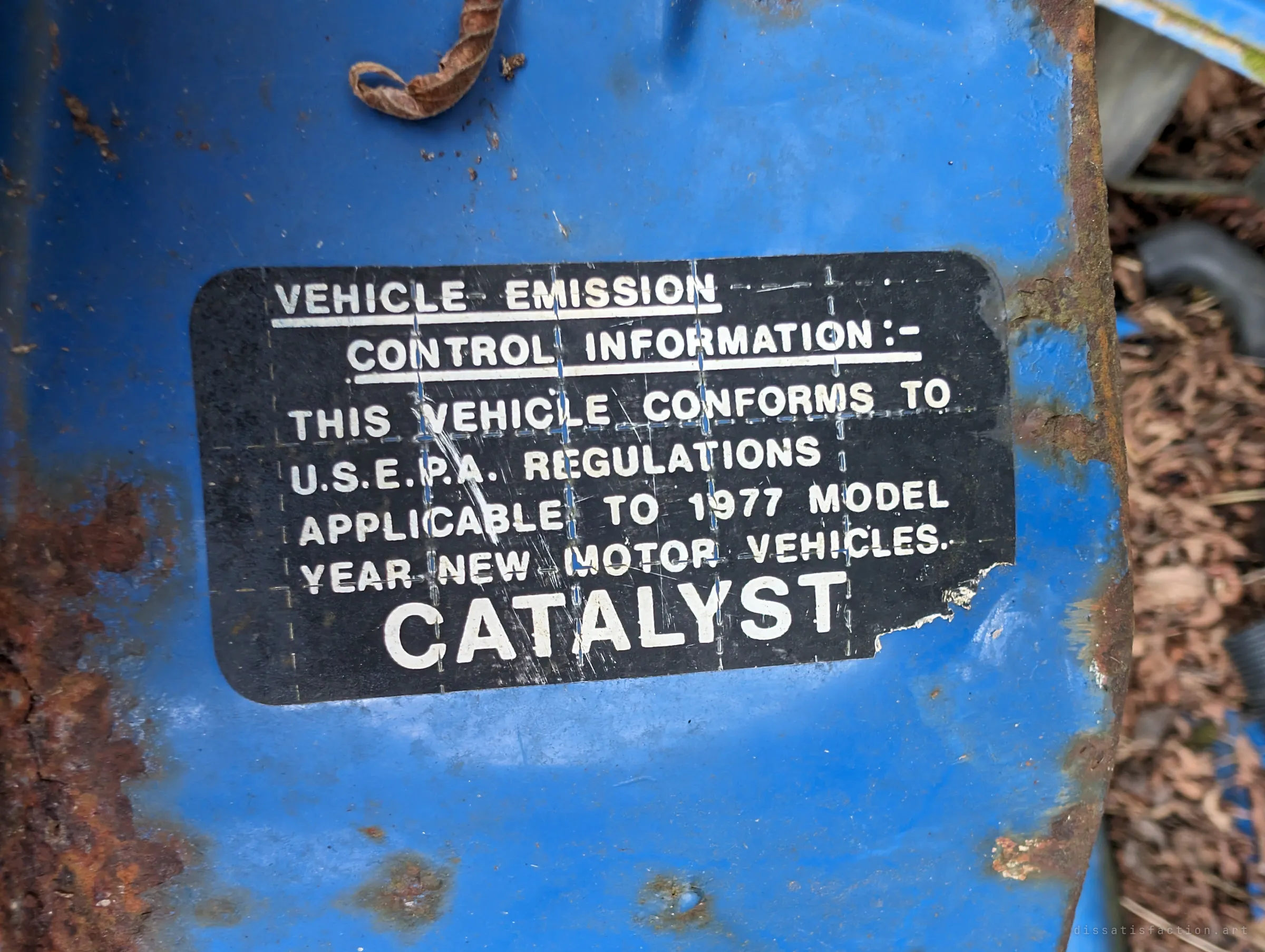 A black factory emissions label on blue metal names the 1977 model year and a catalyst, framed by creeping rust. It dates the wreck to the late seventies.