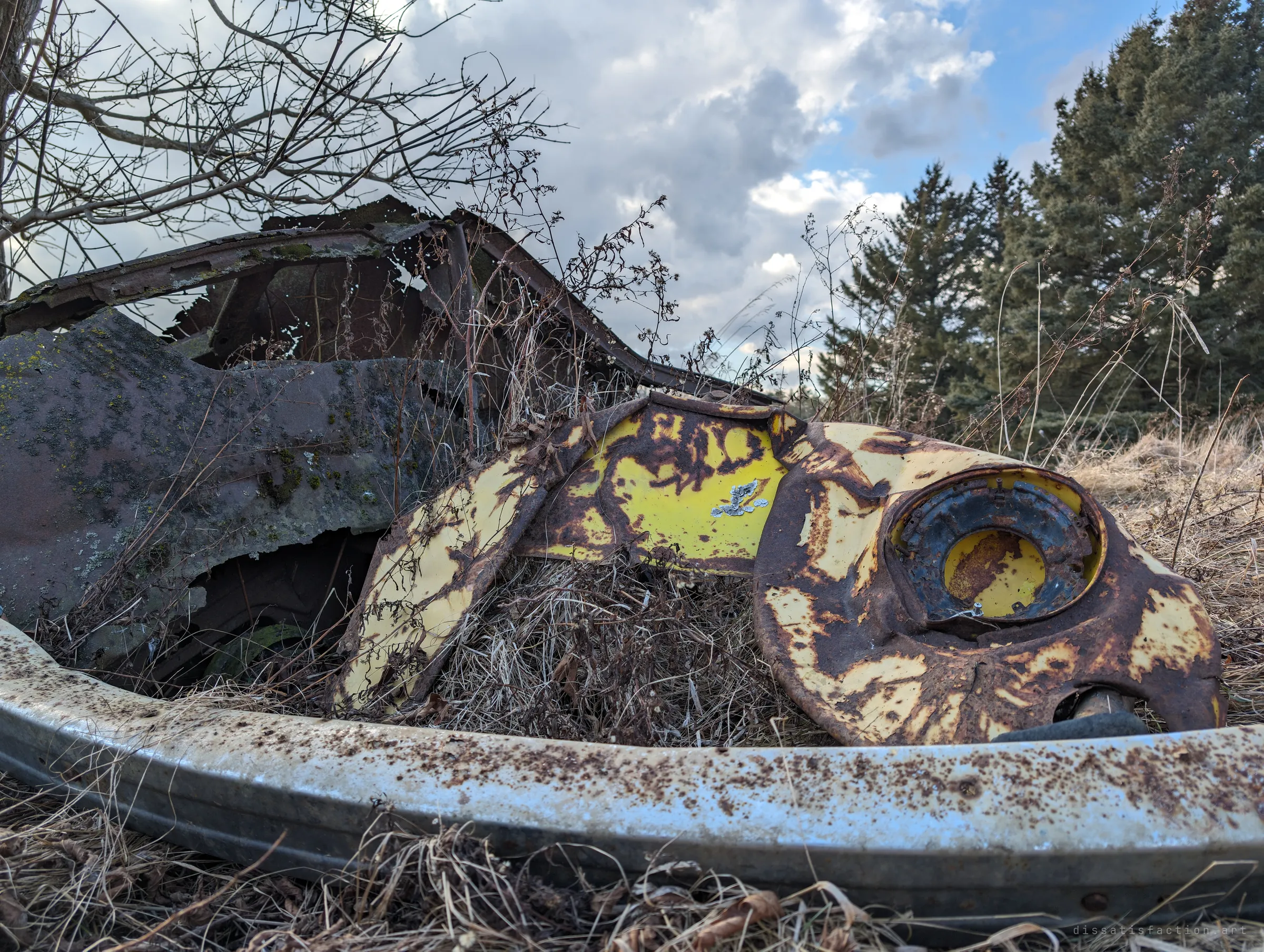 A yellow fender with an empty headlight bowl, blistered with rust, rests against a lichen-covered body panel behind a pitted bumper. Evergreens and a cloudy sky frame the wreck.