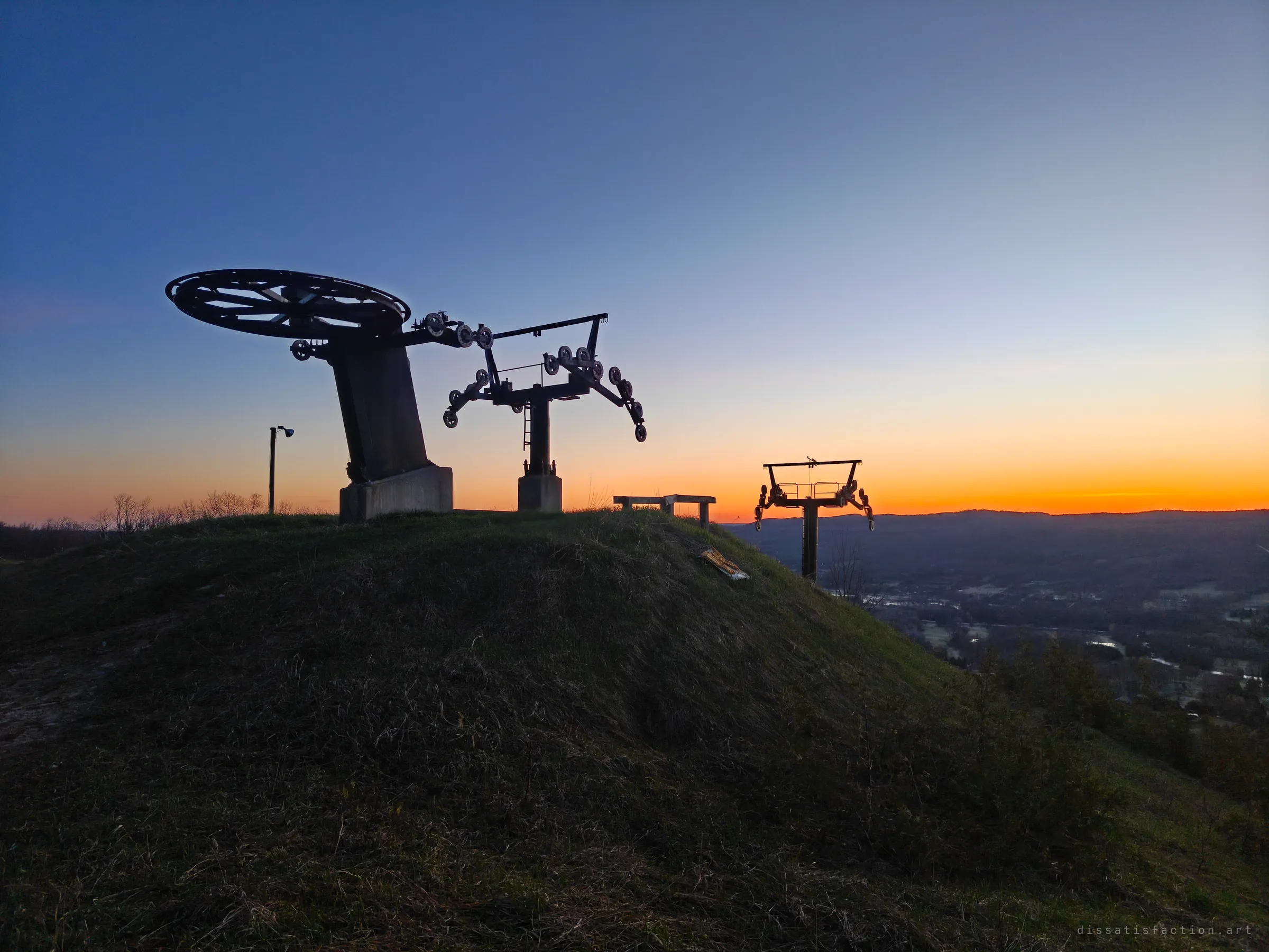 The bull wheel and a lift tower of an abandoned chairlift stand silhouetted on a grassy summit at sunset, with a second tower dropping toward the dark valley. The empty top station shows how the lift simply stopped where it was.