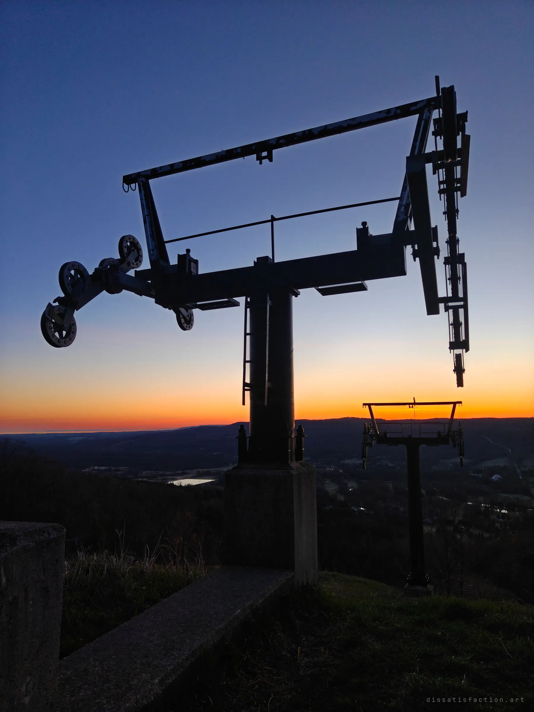 A lift tower's sheave assembly and chipped crossbar loom overhead against an orange dusk sky, with the next tower standing further down the slope. Concrete footings in the foreground mark the old top station.