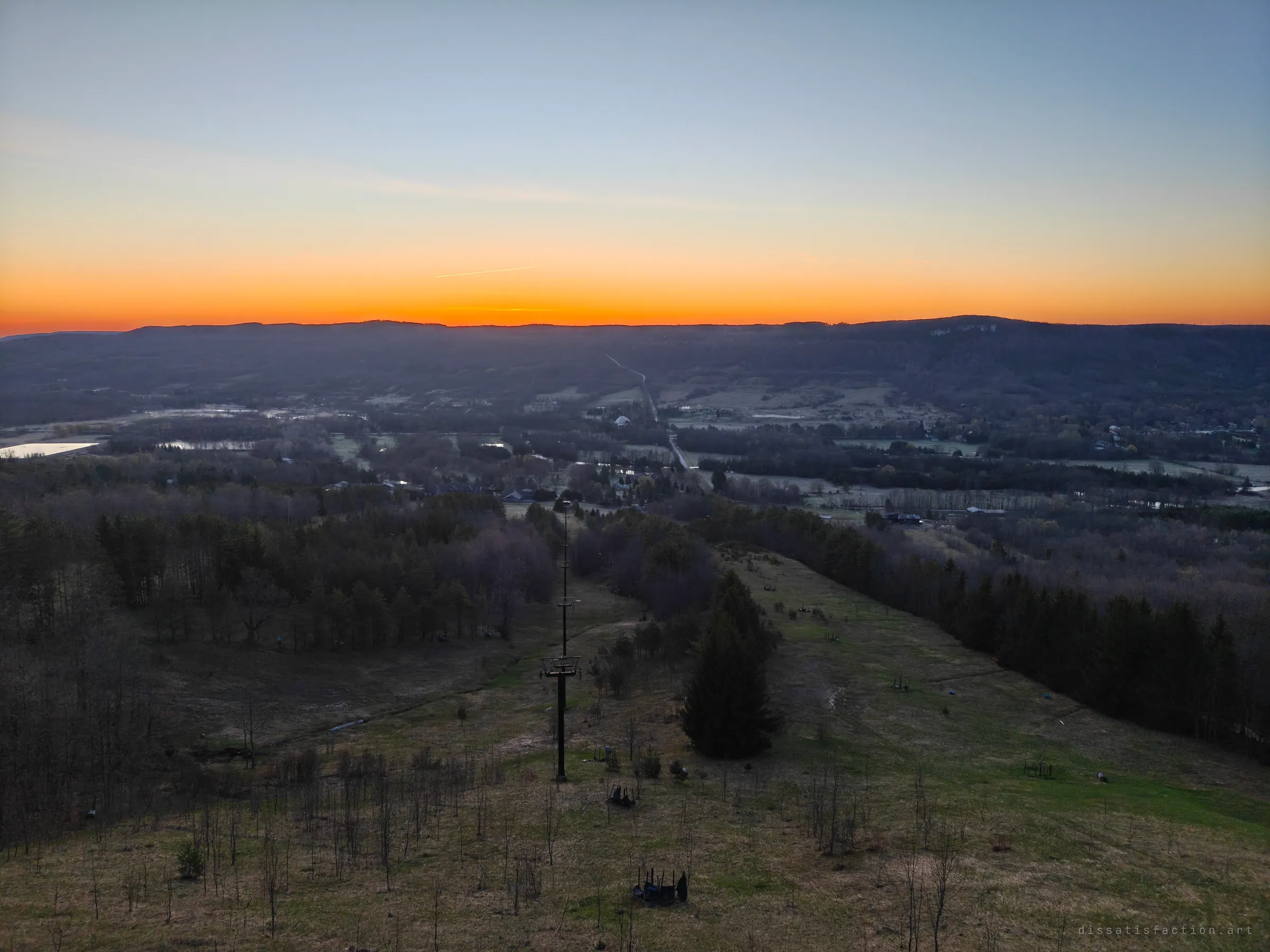 Looking down an overgrown ski run from high above, a single lift tower stands among young trees and scattered snowmaking gear as the sunset glows over the valley. Nature is slowly taking the cleared slope back.