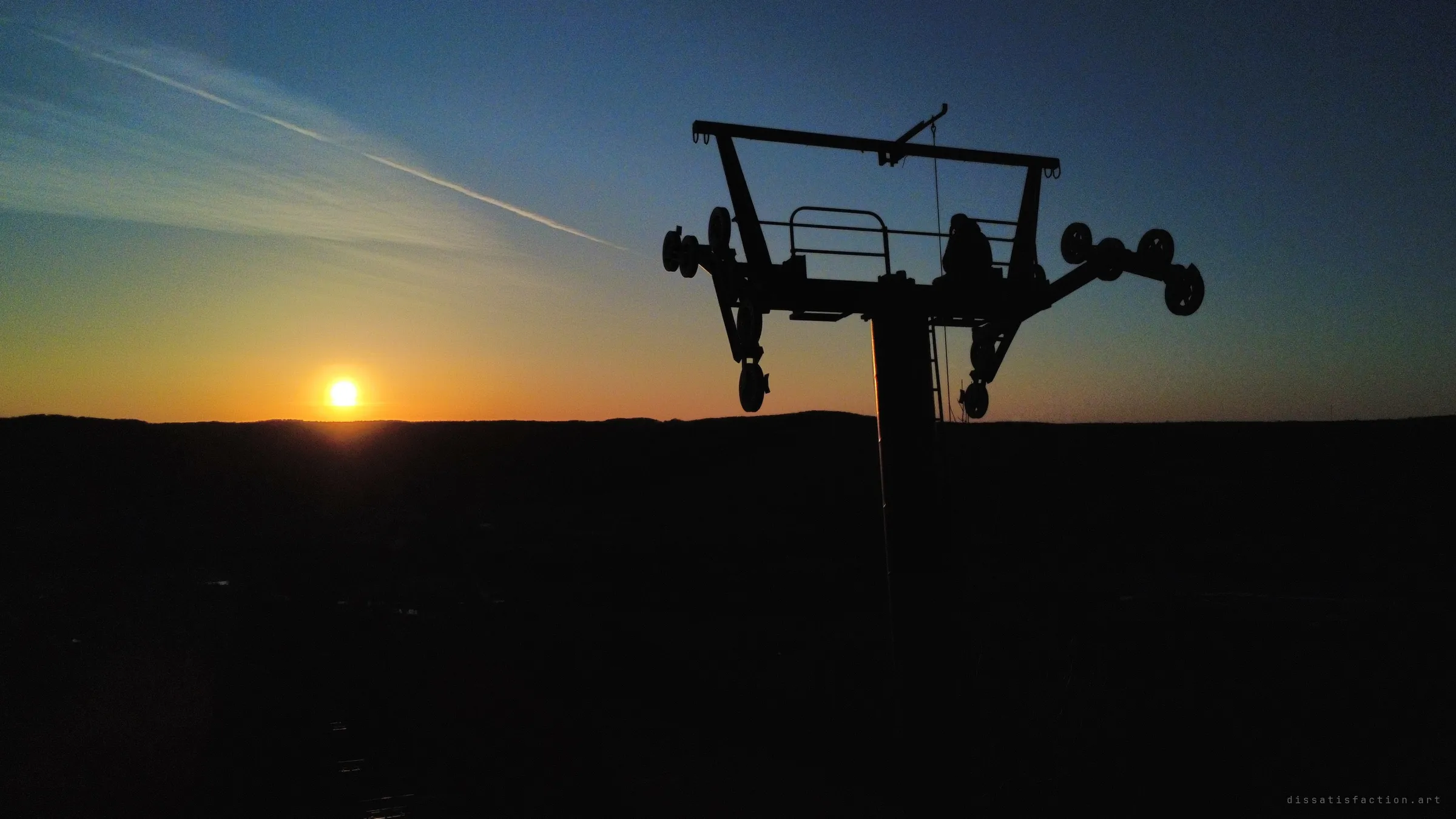 A lone figure sits atop a chairlift tower, silhouetted against the setting sun and a streak of contrail. The drone angle shows how exposed the perch is above the dark hillside.