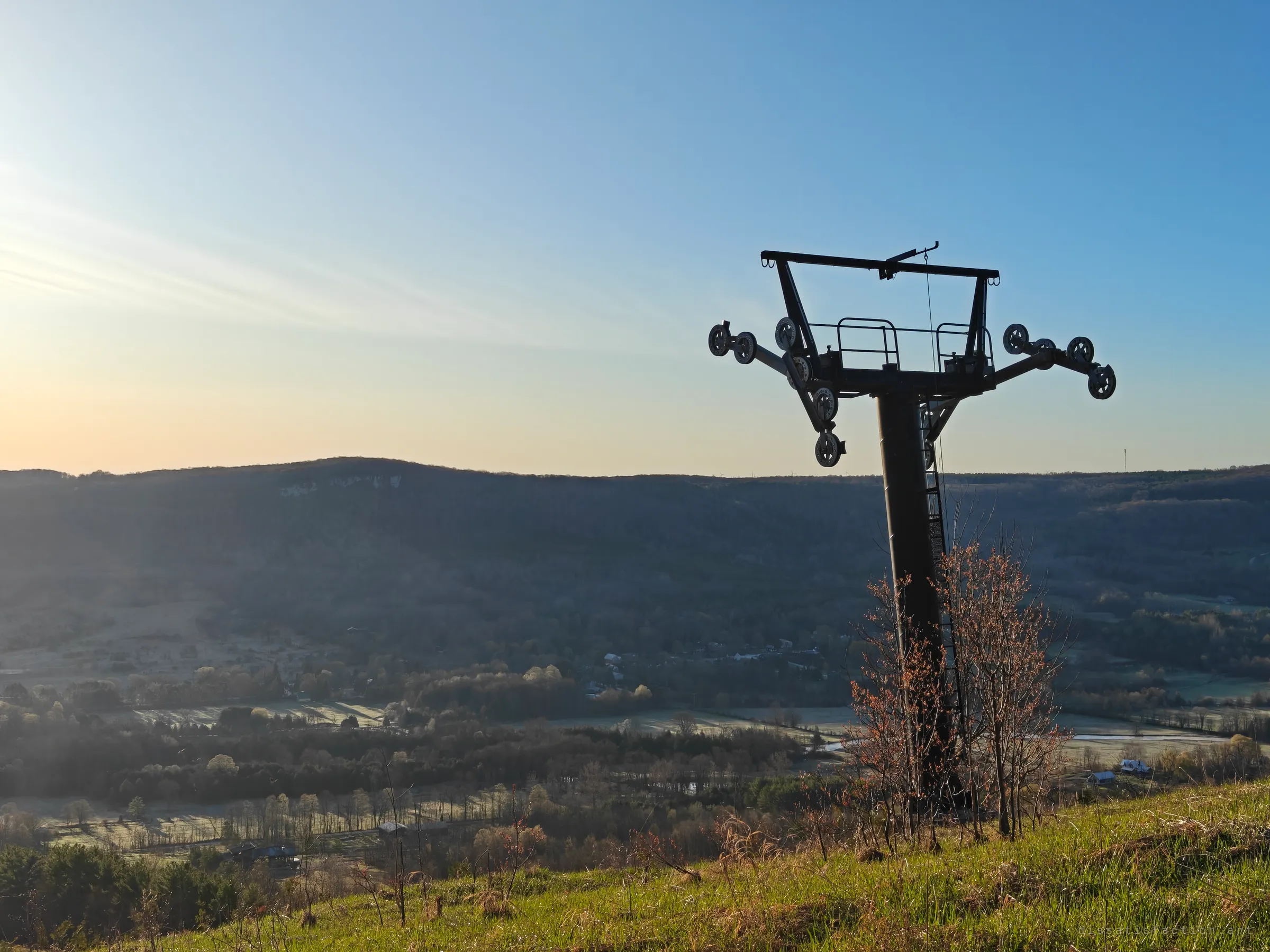 A chairlift tower with its sheave trains rises above bare saplings on a grassy slope, looking across a frosty valley in soft morning light. Brush growing around the base shows years of neglect.