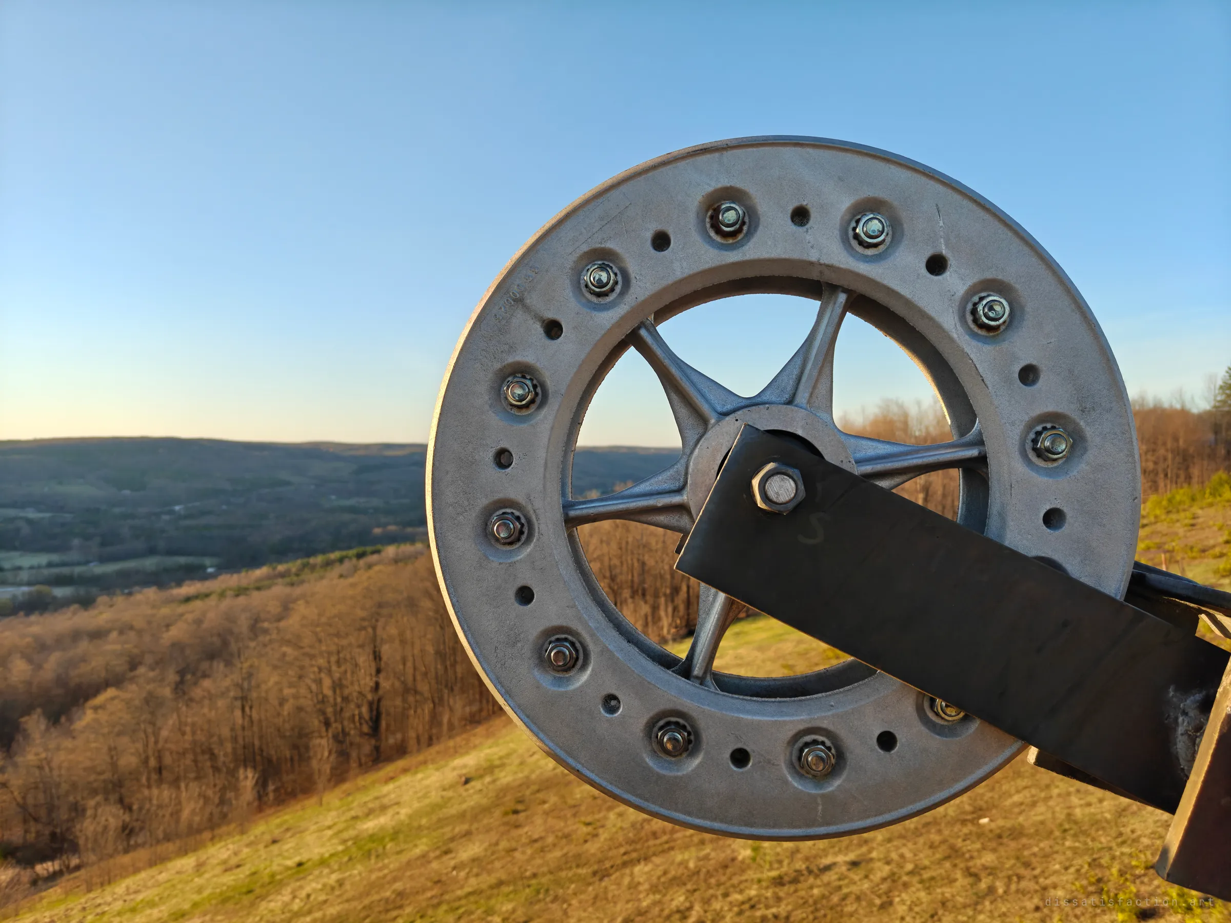 Close-up of a weathered chairlift sheave wheel and its bolted arm, with the forested hillside and valley softly blurred behind. The worn metal shows the scale of the machinery up close.