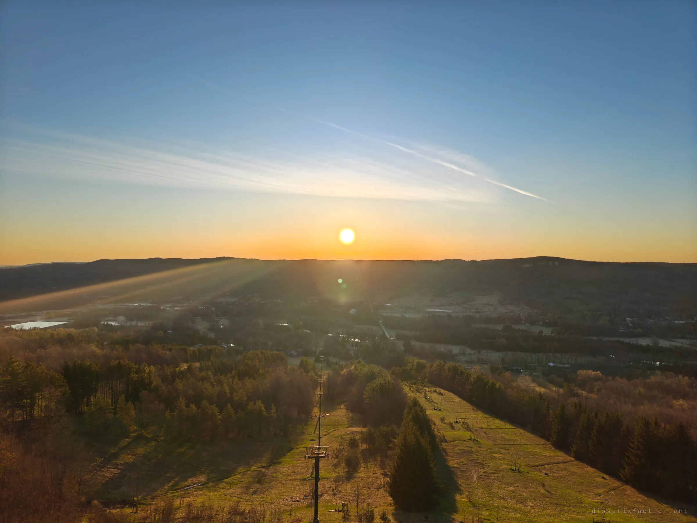 Sunset over a wide valley seen from the top of an overgrown ski run, a lone lift tower running down the grassy slope. It shows how far the abandoned hill looks out over the land it once served.