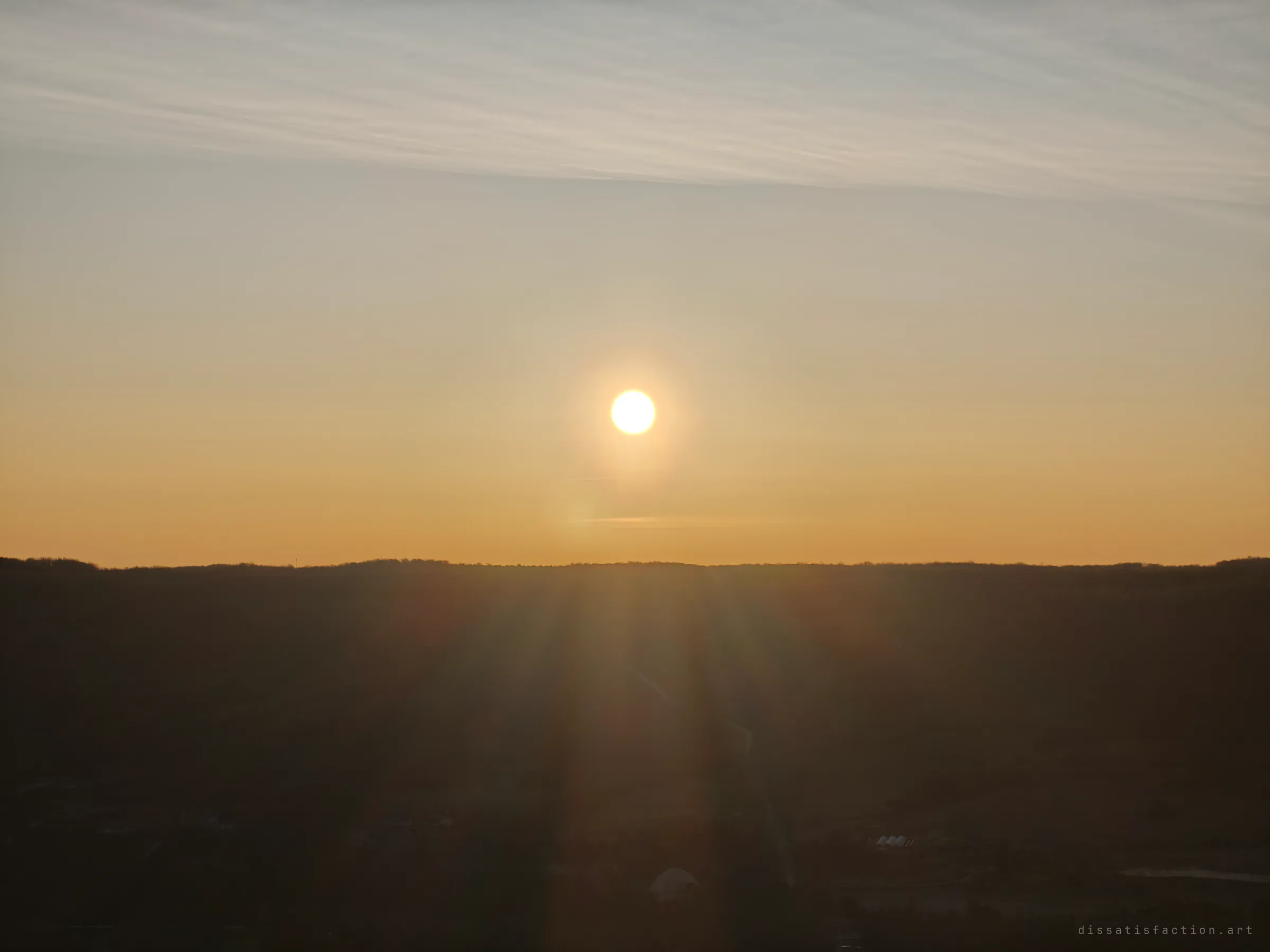 The sun hangs low over a dark ridge, light rays falling across the shadowed valley below. A quiet end-of-day view from the abandoned hilltop.