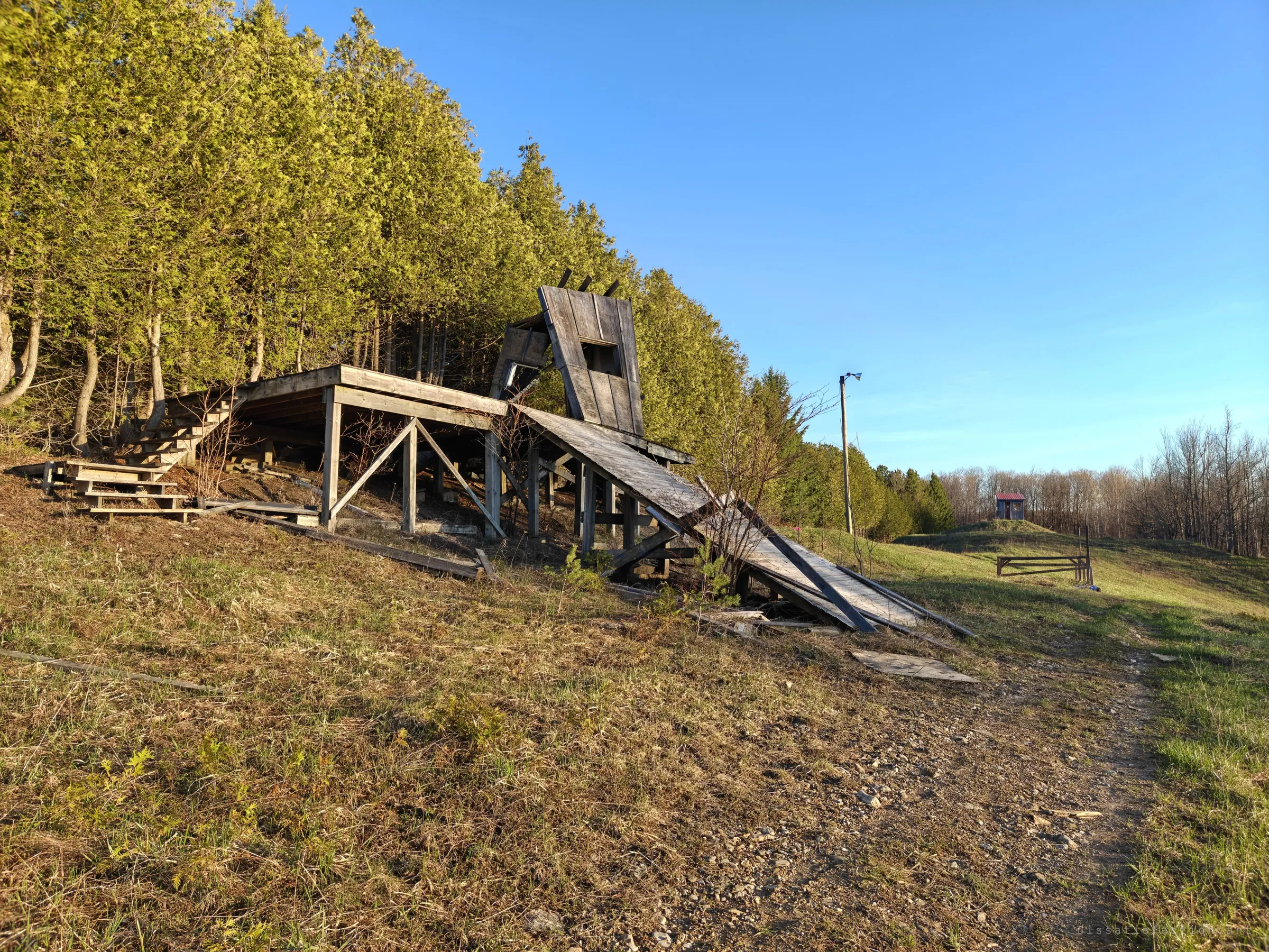 A weathered wooden ramp and deck platform sag into the hillside beneath a wall of cedars, its angled shelter tipping forward. A small red-roofed hut sits on a mound farther along the run.