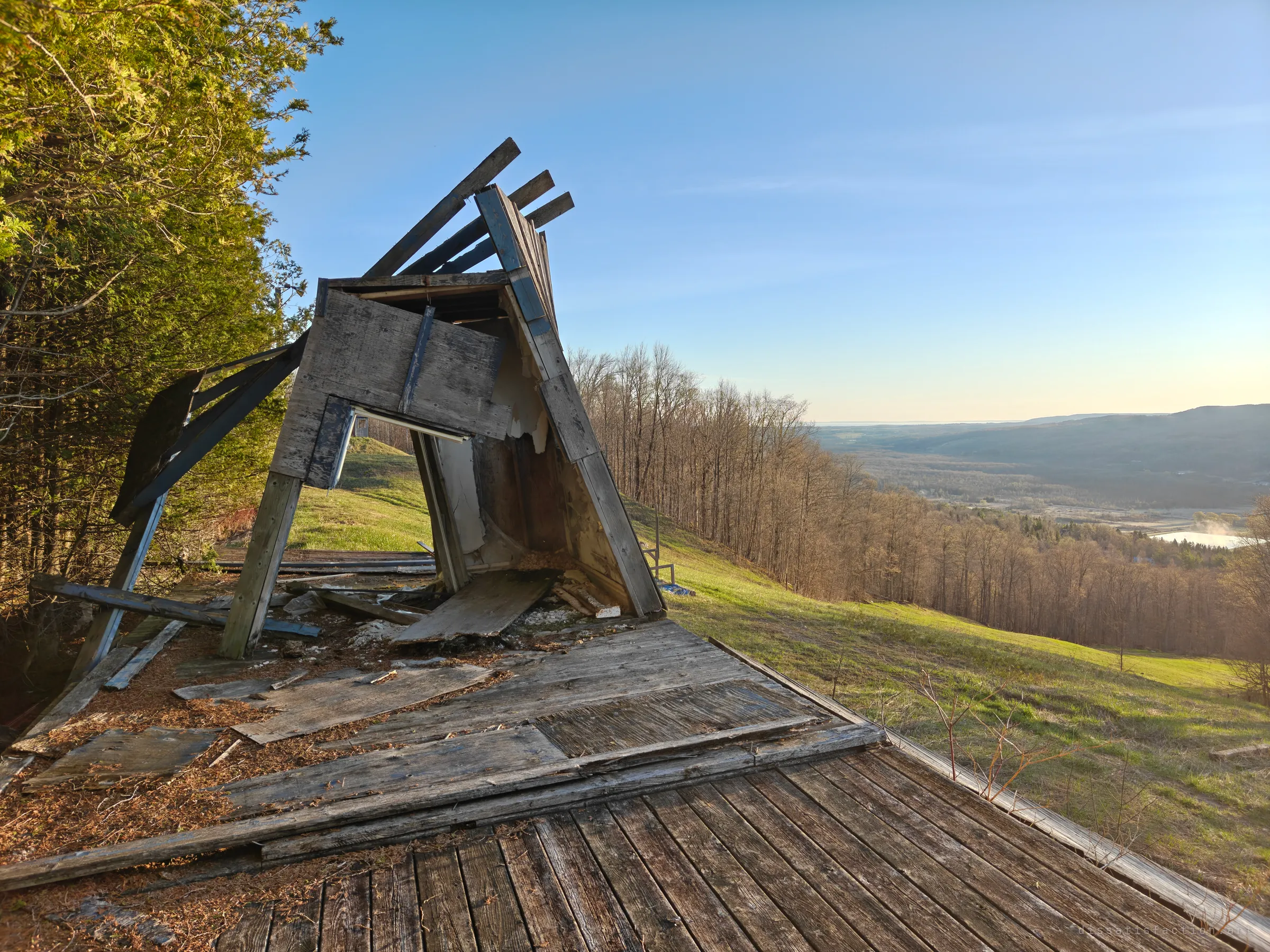 The collapsed wooden shelter at the top of the ramp leans over a buckled plank deck, with the valley opening out beyond the slope. Wind and time have pulled the structure apart board by board.