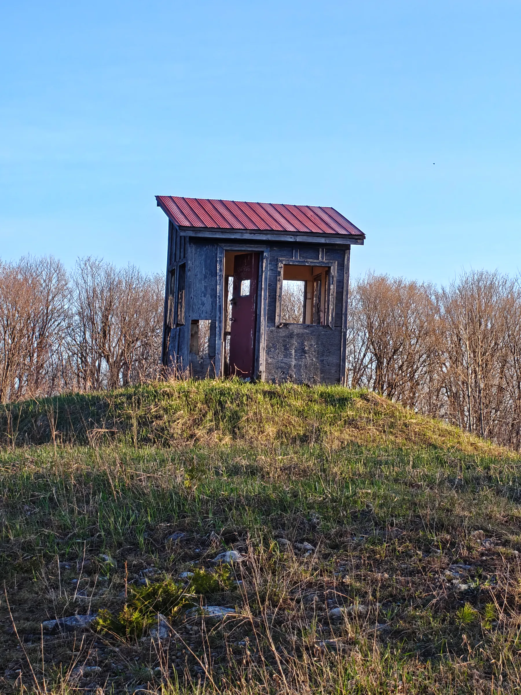 A small grey hut with a red metal roof stands alone on a grassy mound, its maroon door hanging open and window frames empty. The old operator's shack still keeps watch over the run.