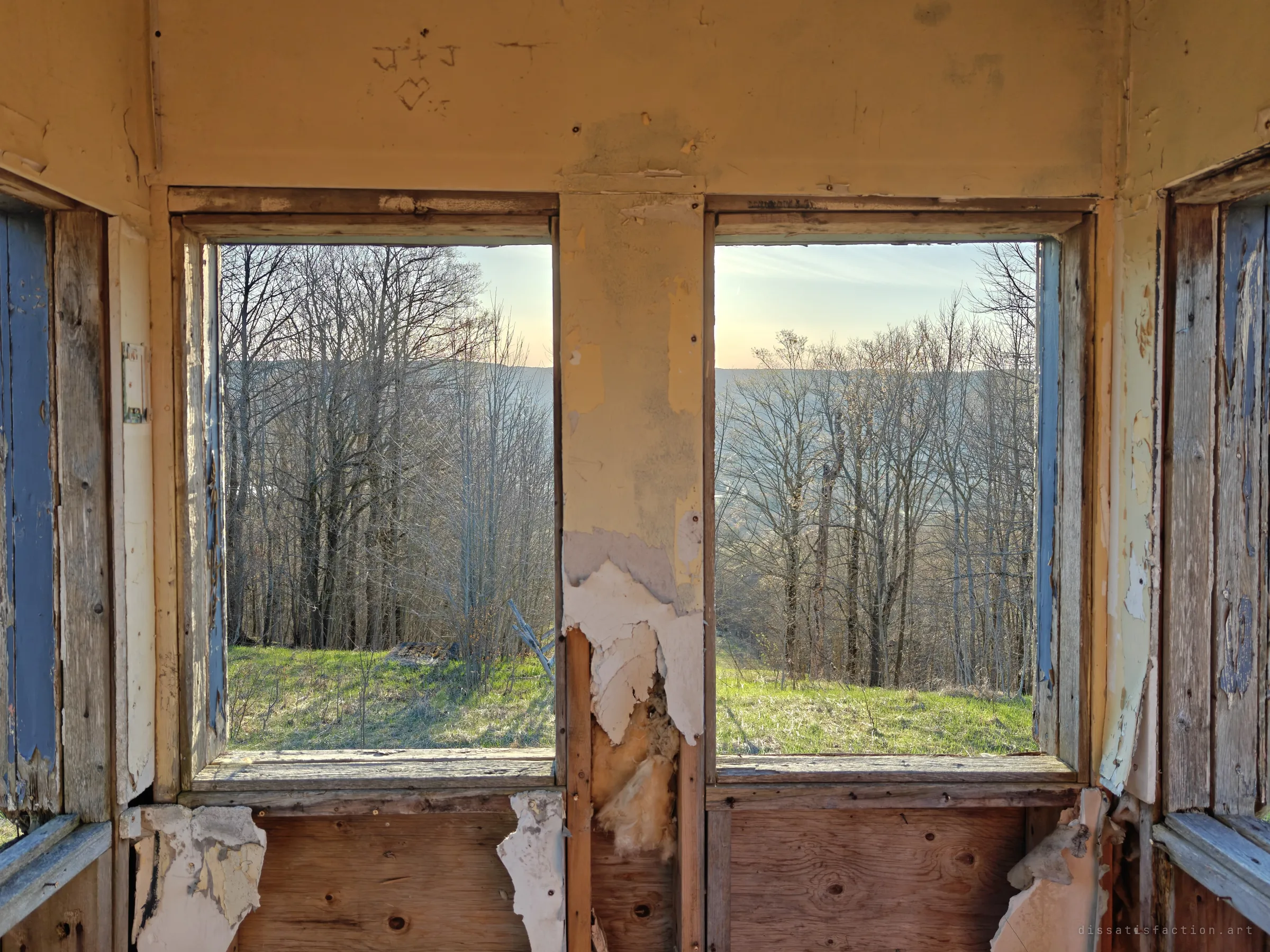 Inside the hut, peeling paint and exposed insulation frame two empty windows looking out onto trees and the fading valley. Carved initials and a heart remain on the wall above.