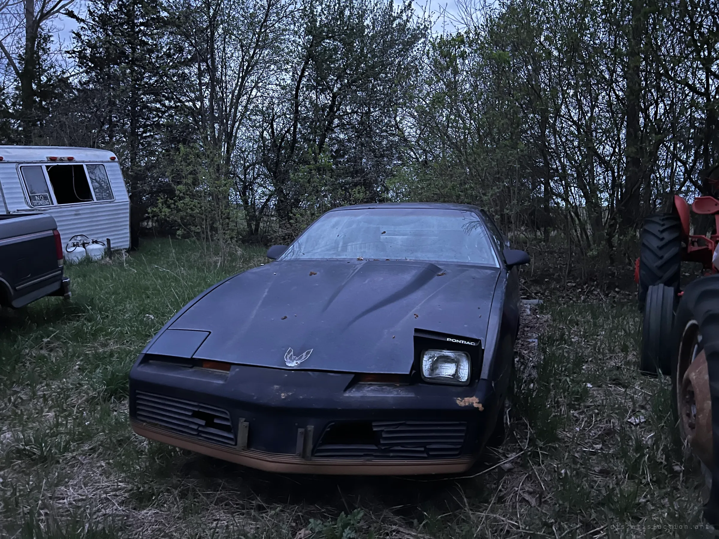 A dusty black 1980s sports coupe sits in long grass at dusk, one pop-up headlight stuck open, with an old camper trailer and a red tractor on either side. It sets the scene of a rural yard where vehicles were left to rot.