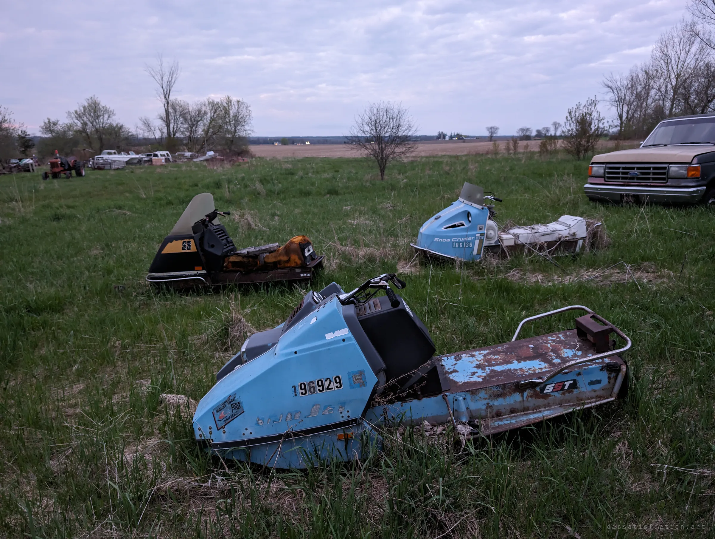 Three vintage snowmobiles lie abandoned in a grassy field, paint faded and seats torn, with a row of derelict cars and a pickup at the edges. The open field shows how far the scrapped machines spread across the property.
