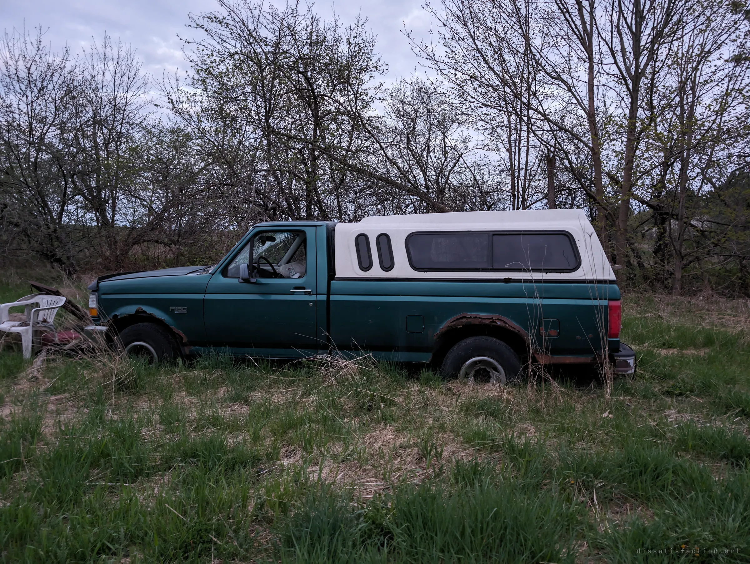 A green full-size pickup with a white cap rests in tall grass beside a plastic lawn chair, its windshield cracked. It reads like it was parked one day and never moved again.