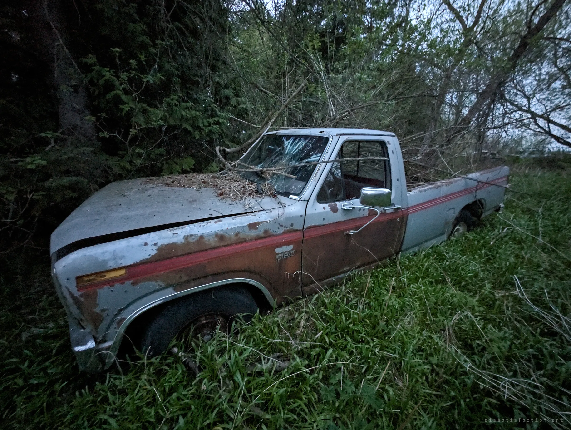 A pale grey and red pickup with a shattered windshield and rust-eaten doors sinks into bright grass beneath overhanging branches. Debris piled on the hood shows years of neglect.
