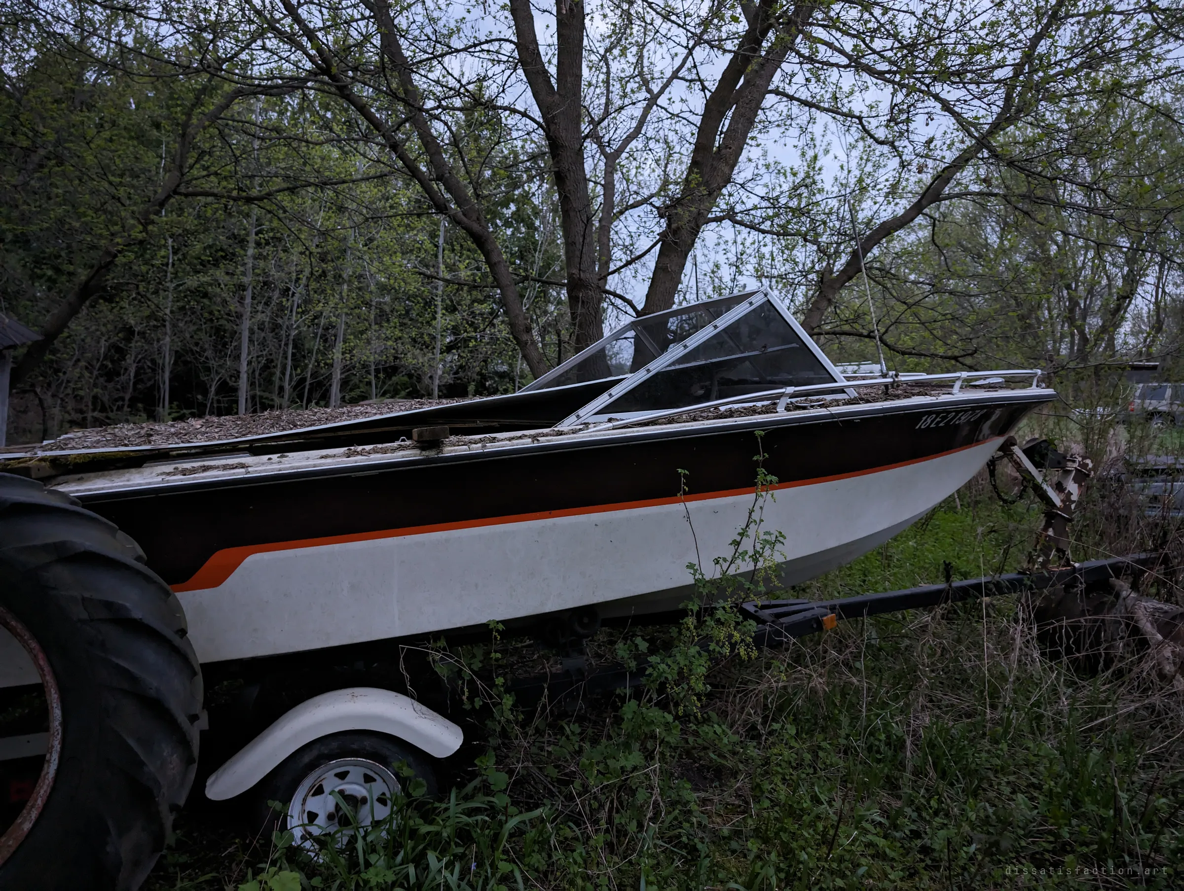 An old runabout boat with a brown and orange stripe rests on a trailer under trees, its deck buried in leaf litter, next to a huge tractor tire. Even the boats were left behind.
