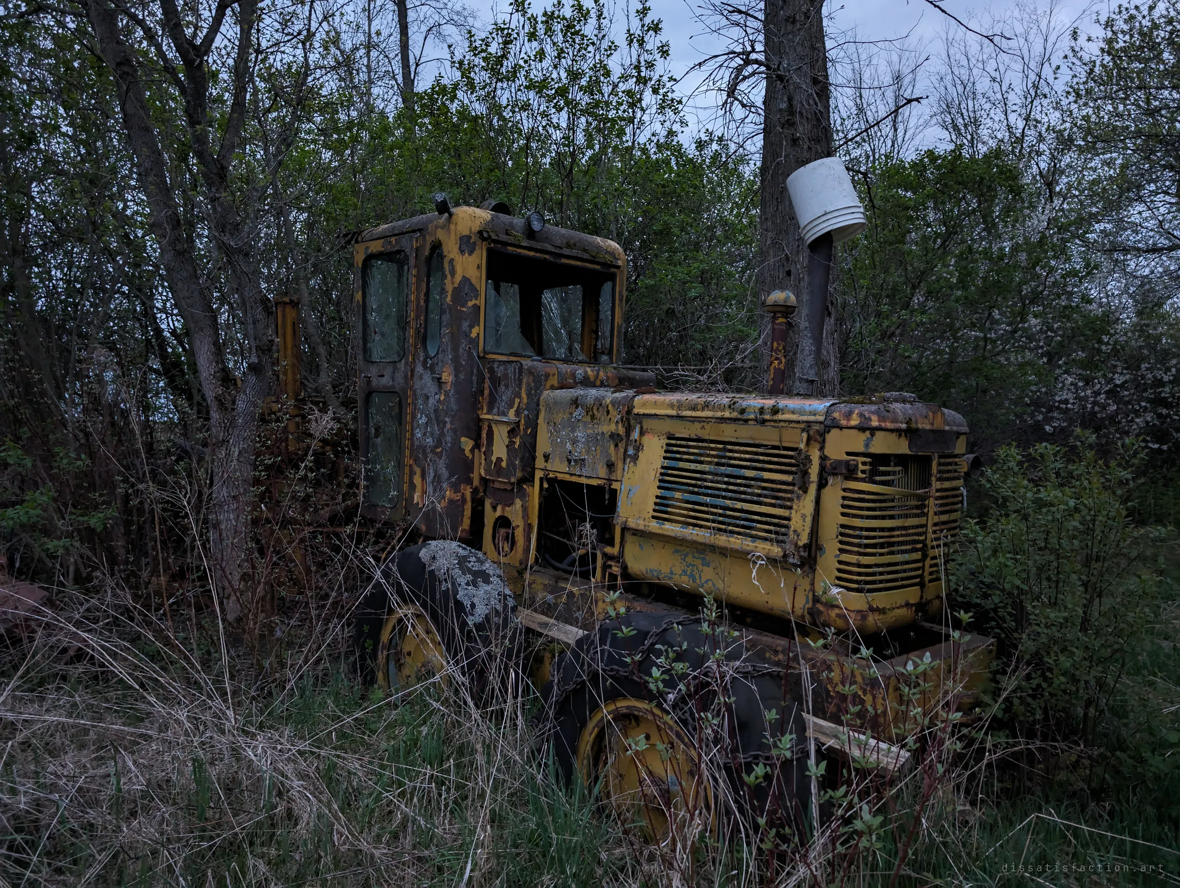 A yellow road grader cab, heavily rusted and patched with lichen, stands in brush with an upturned bucket capping its exhaust stack. Its broken windows and flat tires show decades out of use.