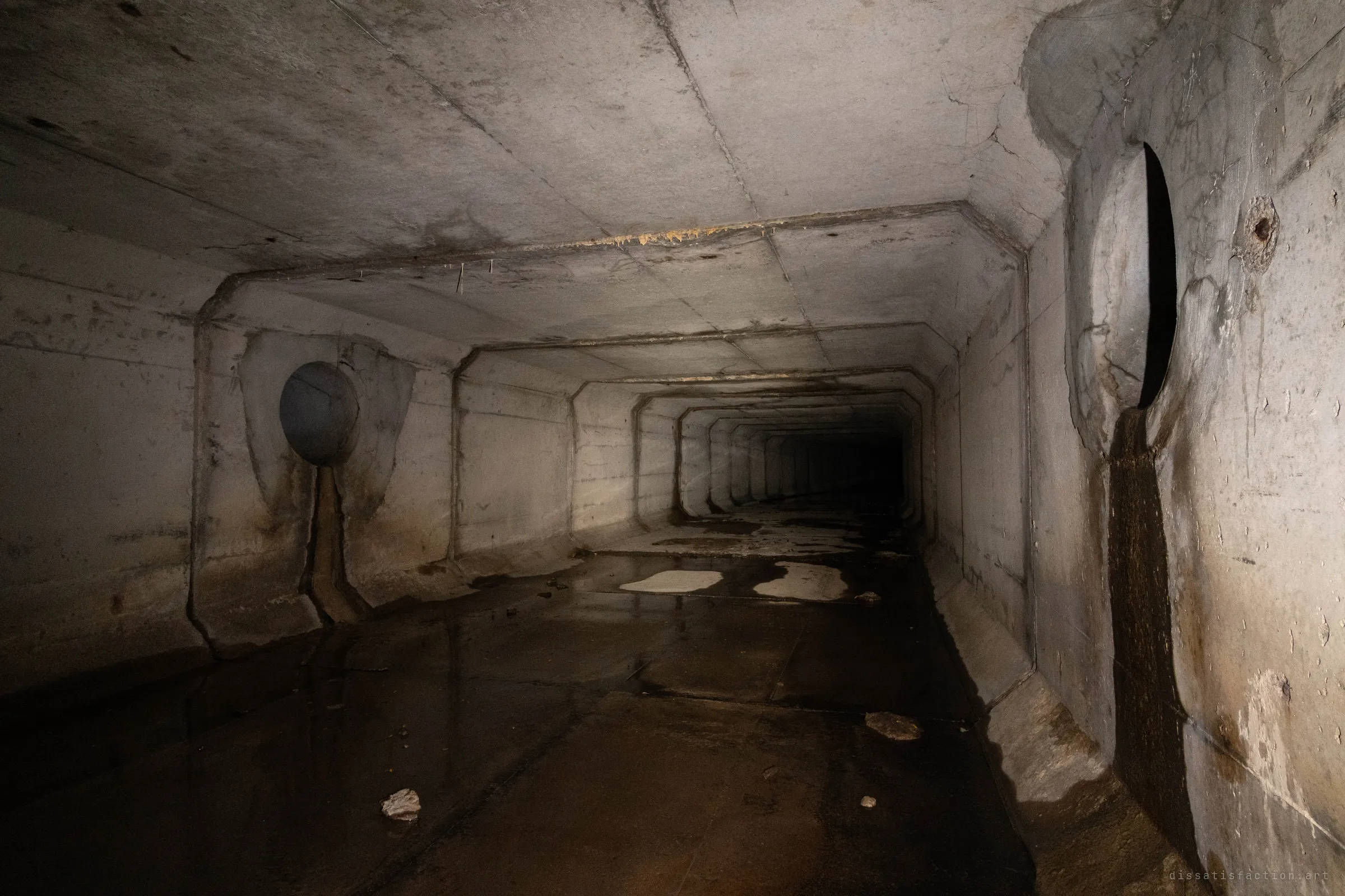 Inside the box culvert, two round side pipes open into the walls, each with a dark streak running down from it to the floor. The chamfered segments repeat into the dark beyond the wet concrete floor.
