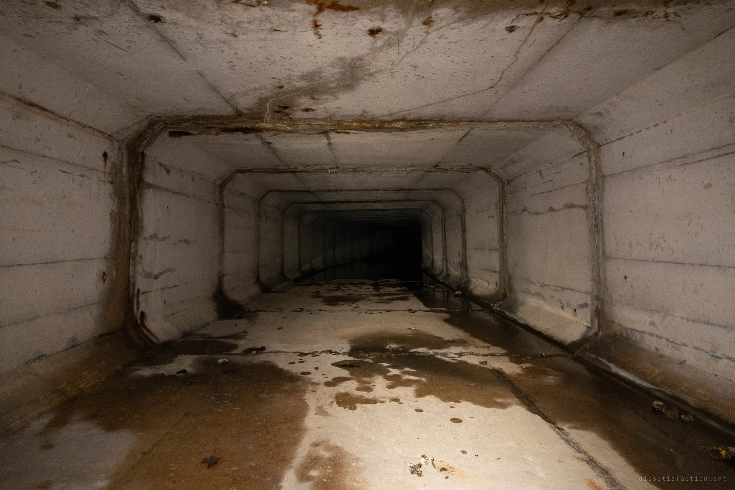 A square concrete box culvert lit by flashlight, its chamfered segment frames stepping away into total darkness. Rust stains on the ceiling and puddles of standing water on the floor show the tunnel still carries water.