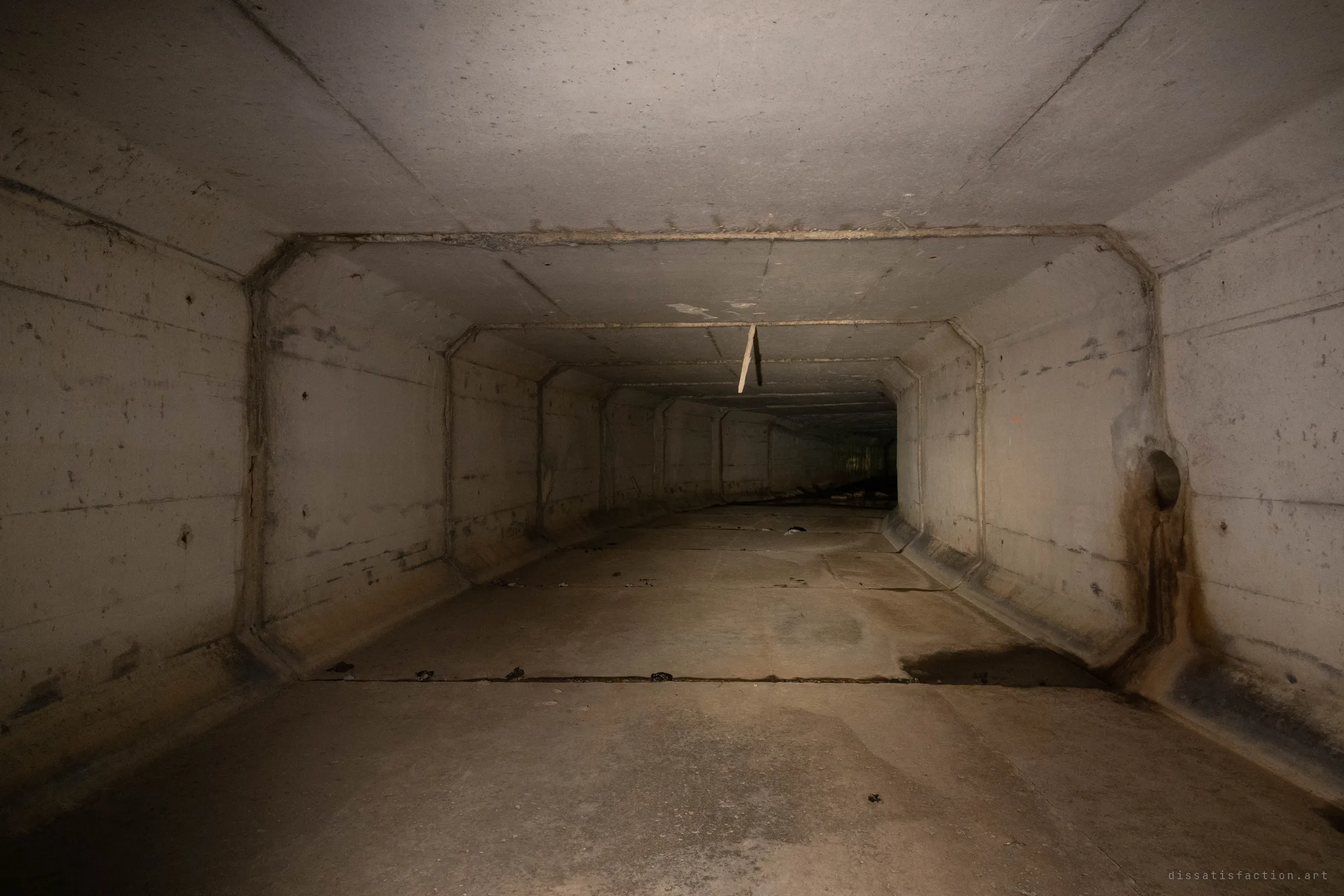 A long, dry stretch of the concrete box culvert running straight into the dark, with a broken stick hanging from a ceiling joint. A rust-streaked inlet pipe opens through the right wall above a damp patch on the floor.