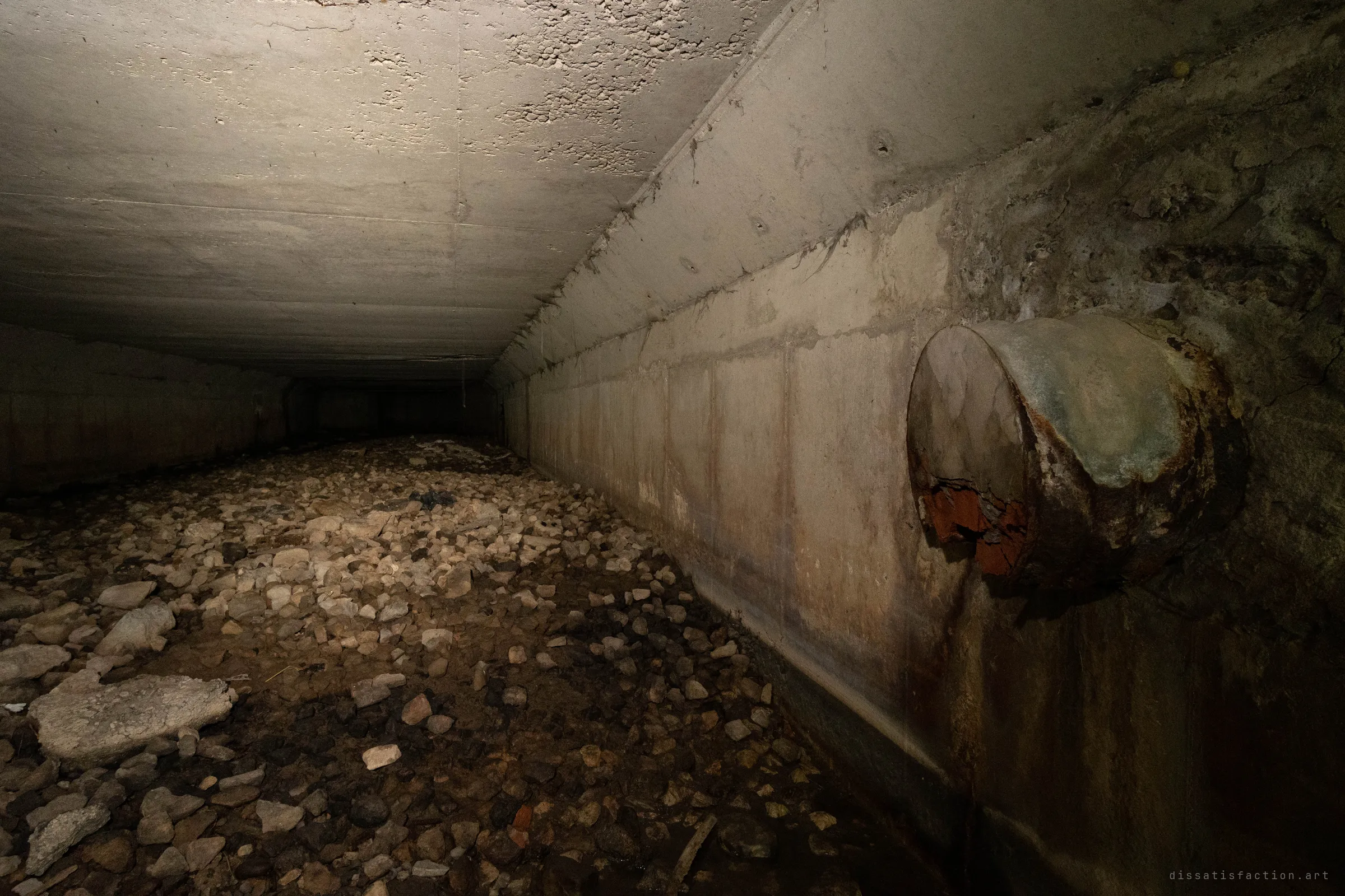 A low concrete box culvert runs into darkness over a bed of loose rubble and pale stones, lit warm from the left. A rusted, torn pipe juts out of the right wall above long streaks of rust and water staining.